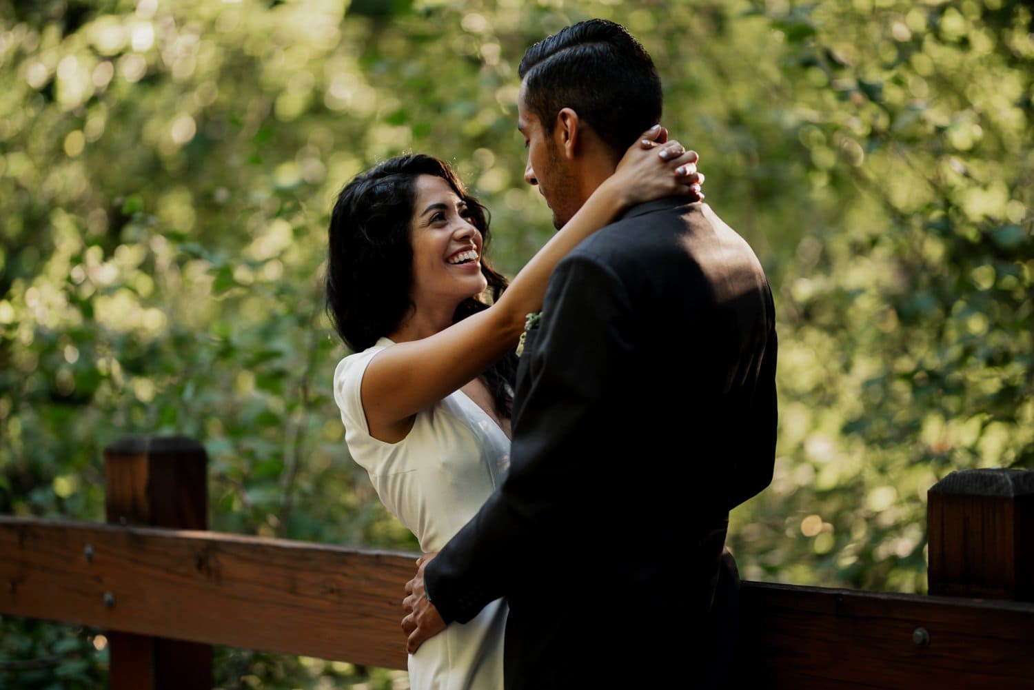 A couple embraces and smiles on a wooden bridge surrounded by lush greenery at Leach Botanical Garden on their wedding day.
