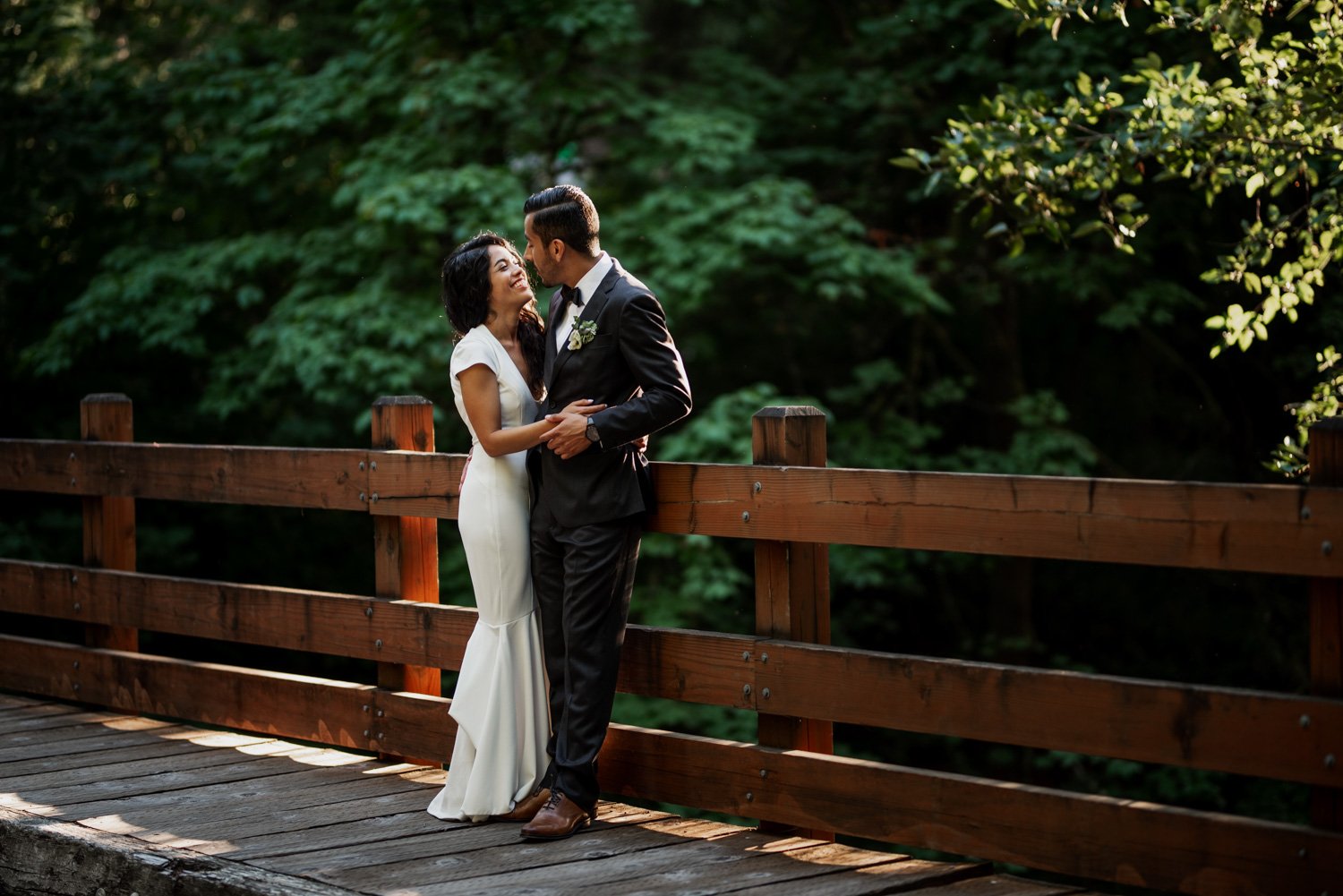 A bride and groom embrace on a wooden bridge at their wedding in Leach Botanical Garden, surrounded by lush greenery.