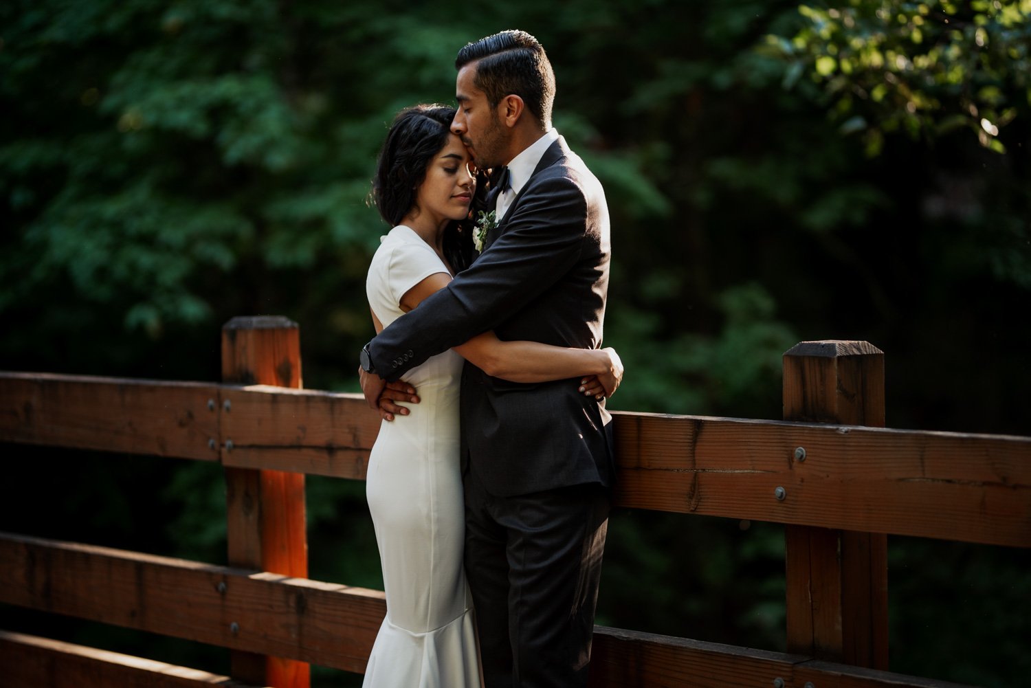 A couple embraces on a wooden bridge at Leach Botanical Garden, surrounded by lush forest. They are dressed in formal attire, adding elegance to the romantic wedding setting.