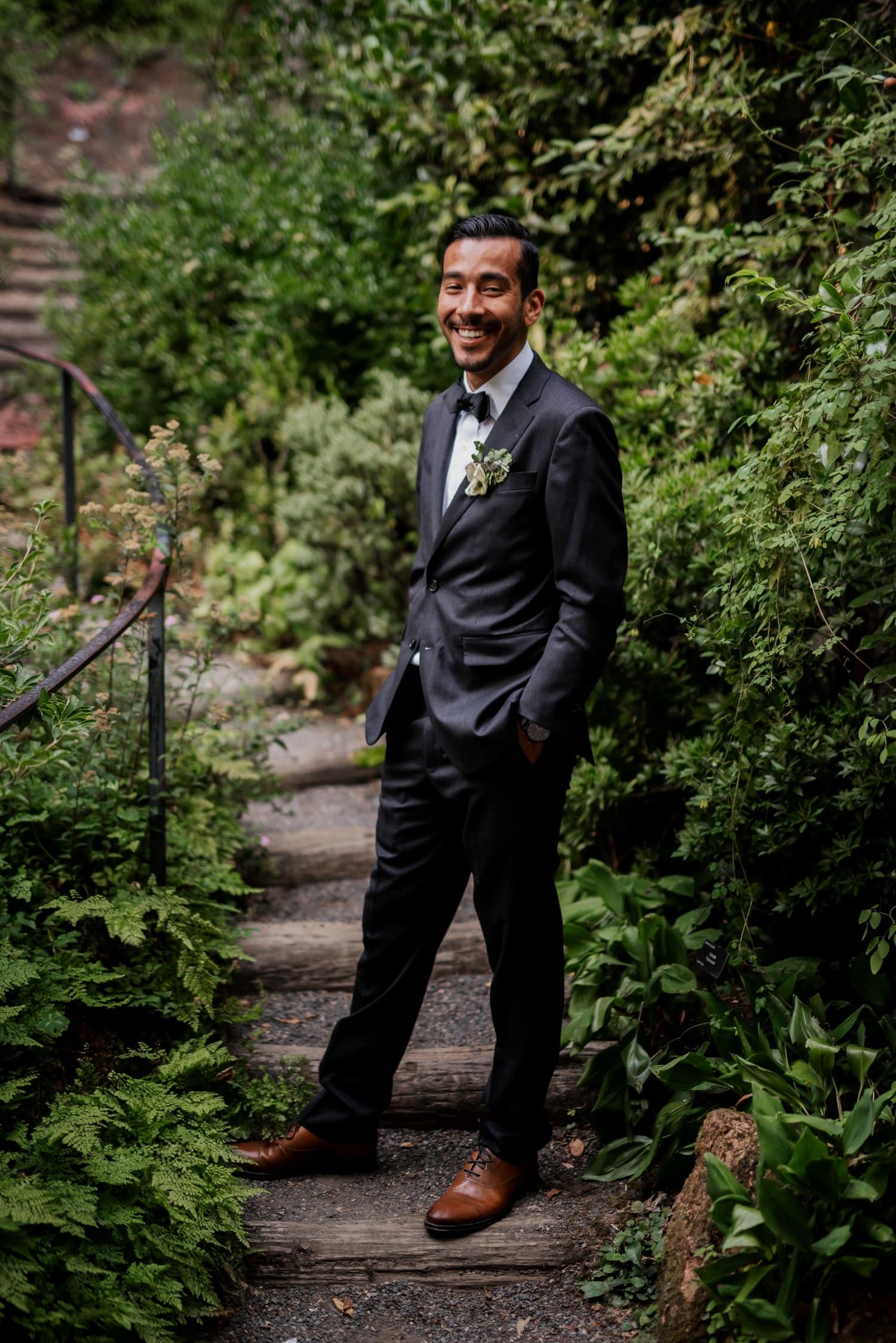 A smiling man in a suit stands on a garden pathway at Leach Botanical Garden, surrounded by lush greenery.