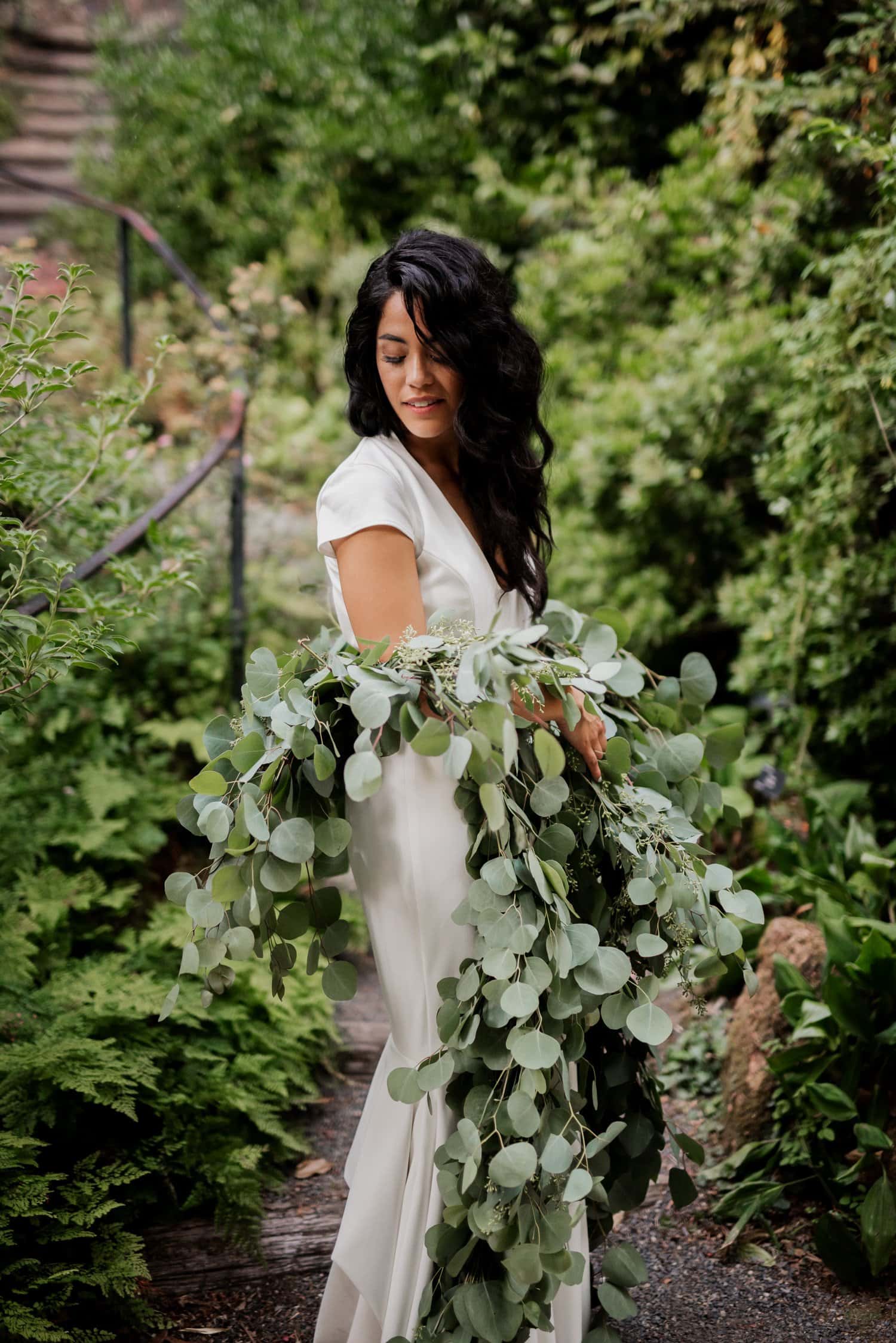 A woman in a white dress holds lush greenery while posing on a garden path surrounded by plants at Leach Botanical Garden.