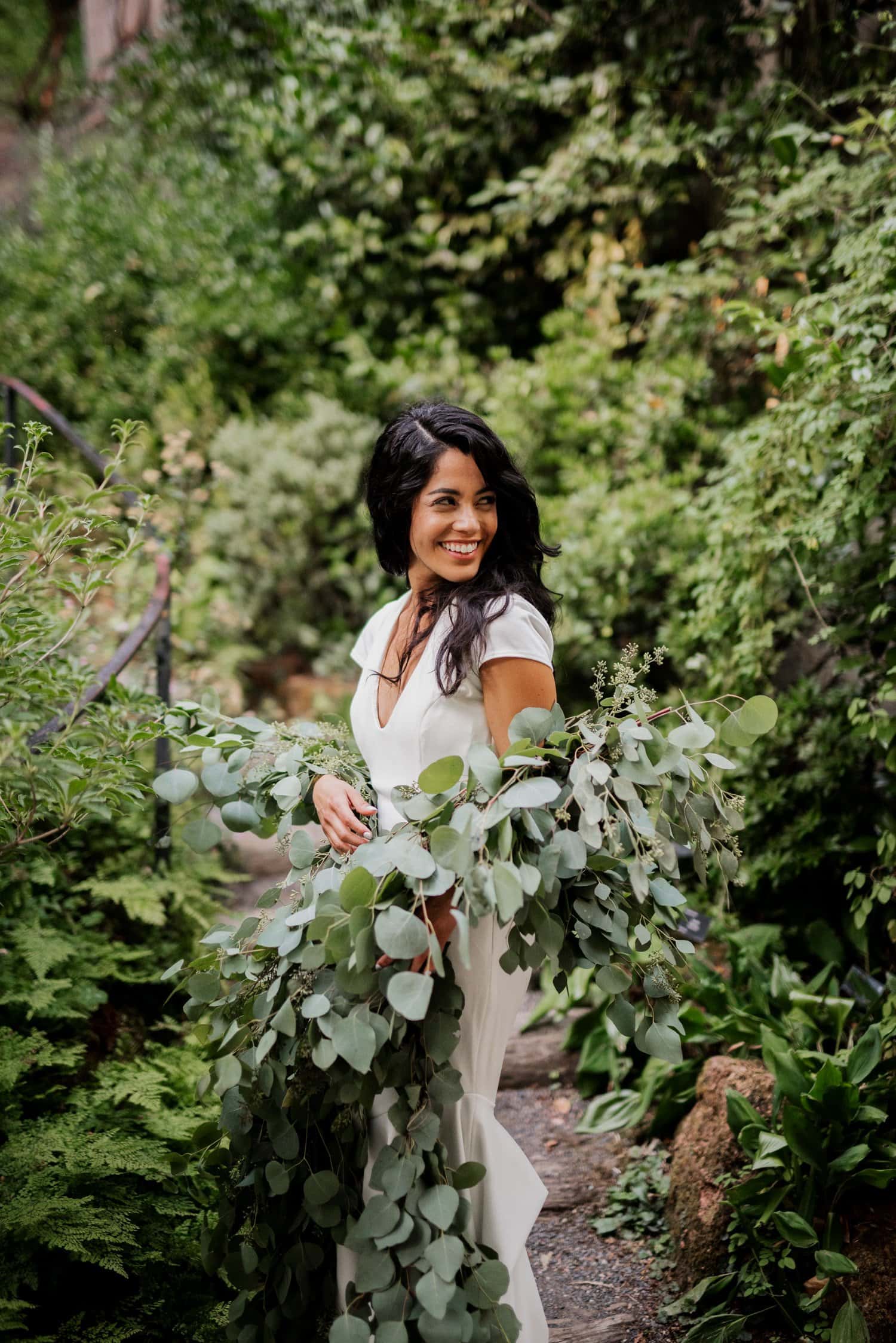 A woman in a white dress smiles while holding a bouquet of green plants, with colorful leaves and plants around her at Leach Botanical Garden.