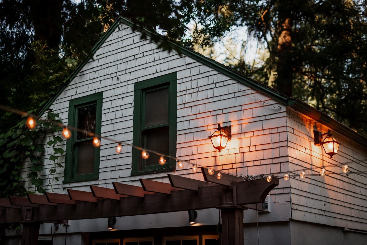 A house with green window frames, string lights, and a wooden pergola surrounded by trees at dusk.