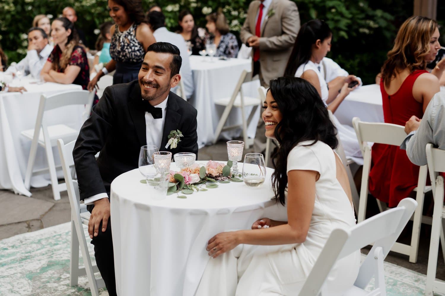 A couple smiles as they sit at a table decorated with flowers and candles during their wedding reception at Leach Botanical Garden.