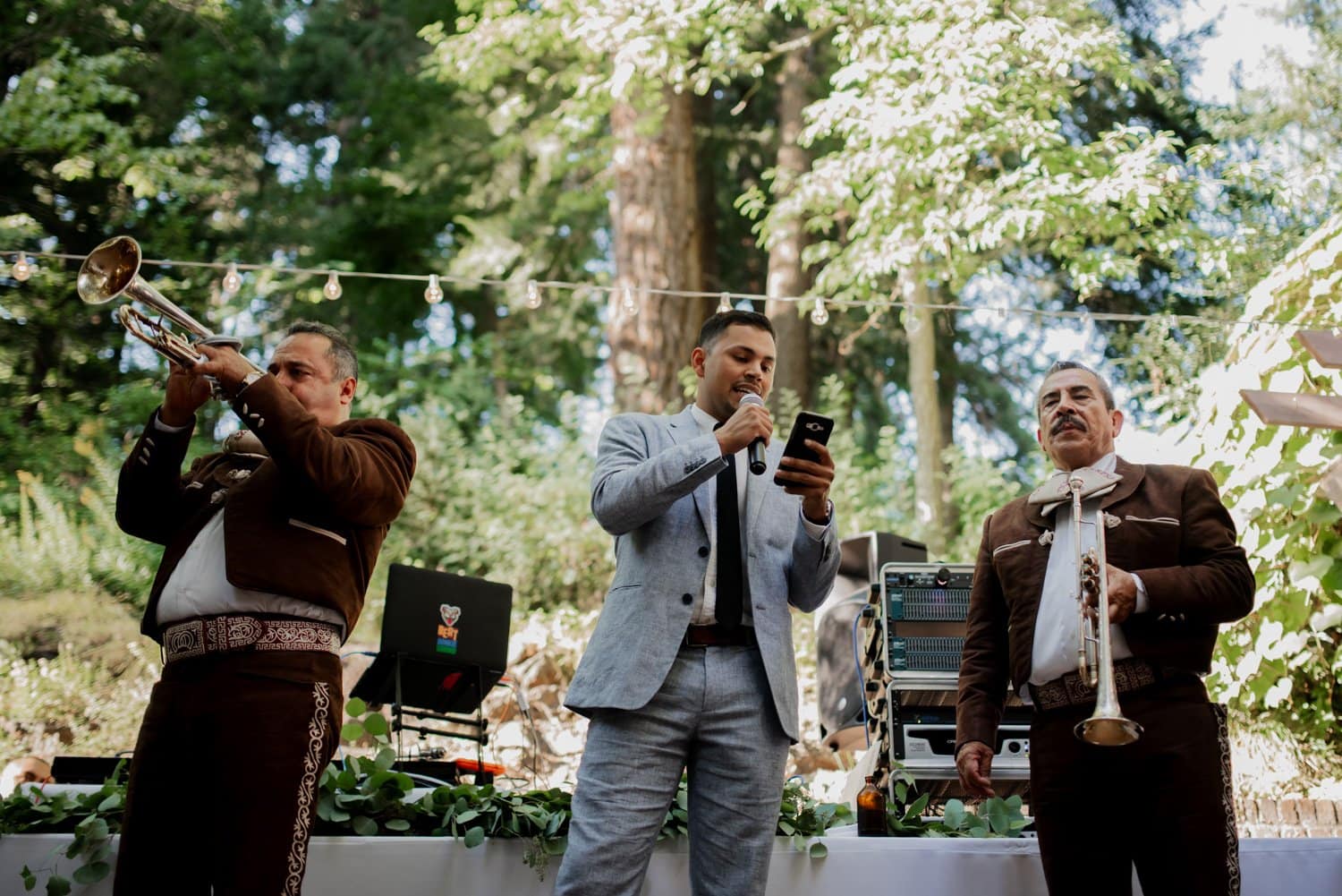 Three musicians perform at a Leach Botanical Garden wedding, with one singer and two trumpet players creating an enchanting outdoor atmosphere. They are surrounded by trees and illuminated by twinkling decorative lights.