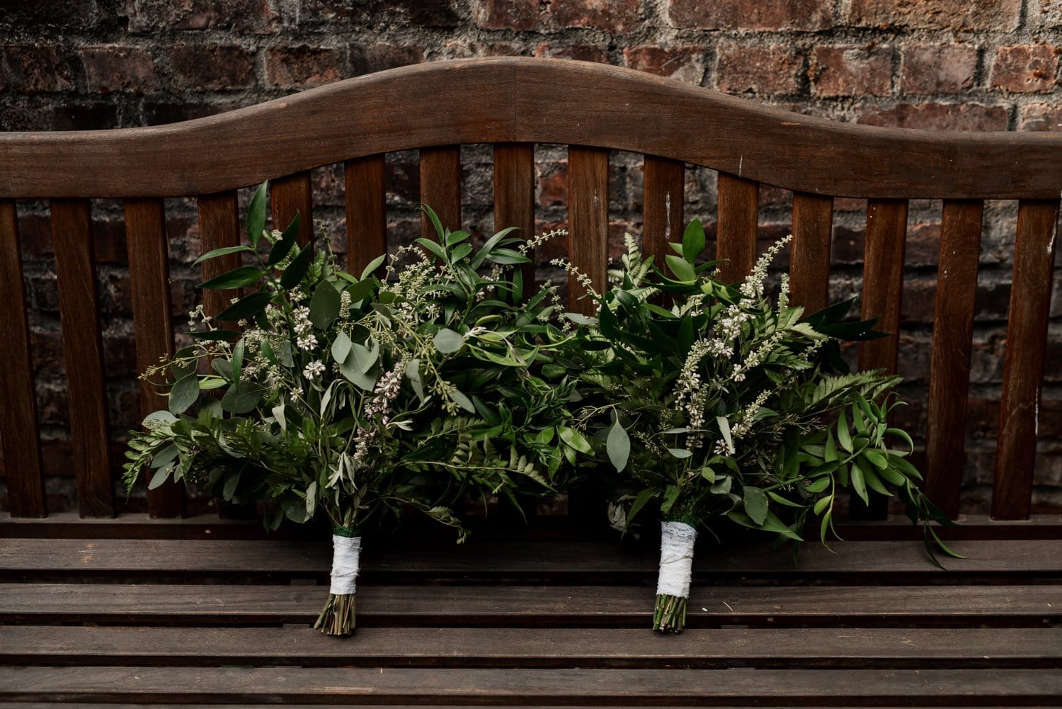 Two green leafy bouquets with white accents on a wooden bench, with a brick wall backdrop at Leach Botanical Garden.