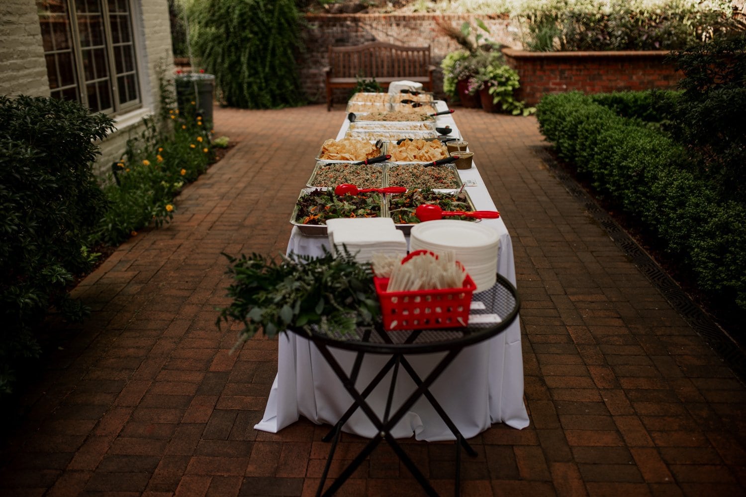 Buffet table with a variety of dishes and plates arranged outdoors on a brick patio, surrounded by lush green plants and trees.
