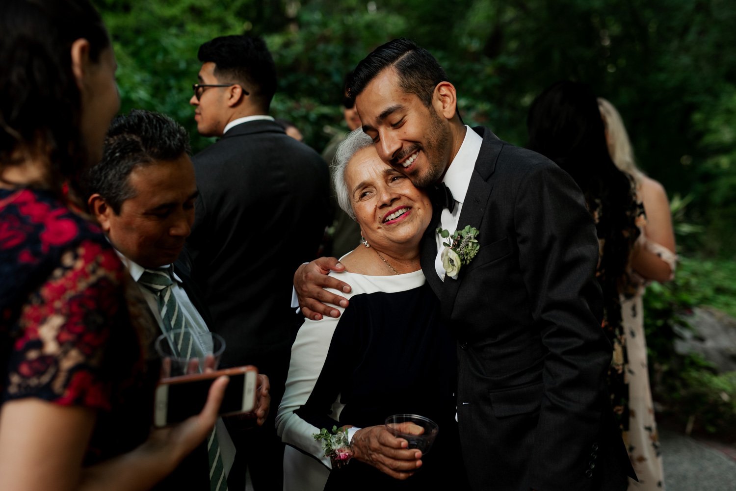 A man in a suit embraces a woman in a formal dress at Leach Botanical Garden, surrounded by elegantly dressed guests and lush greenery.