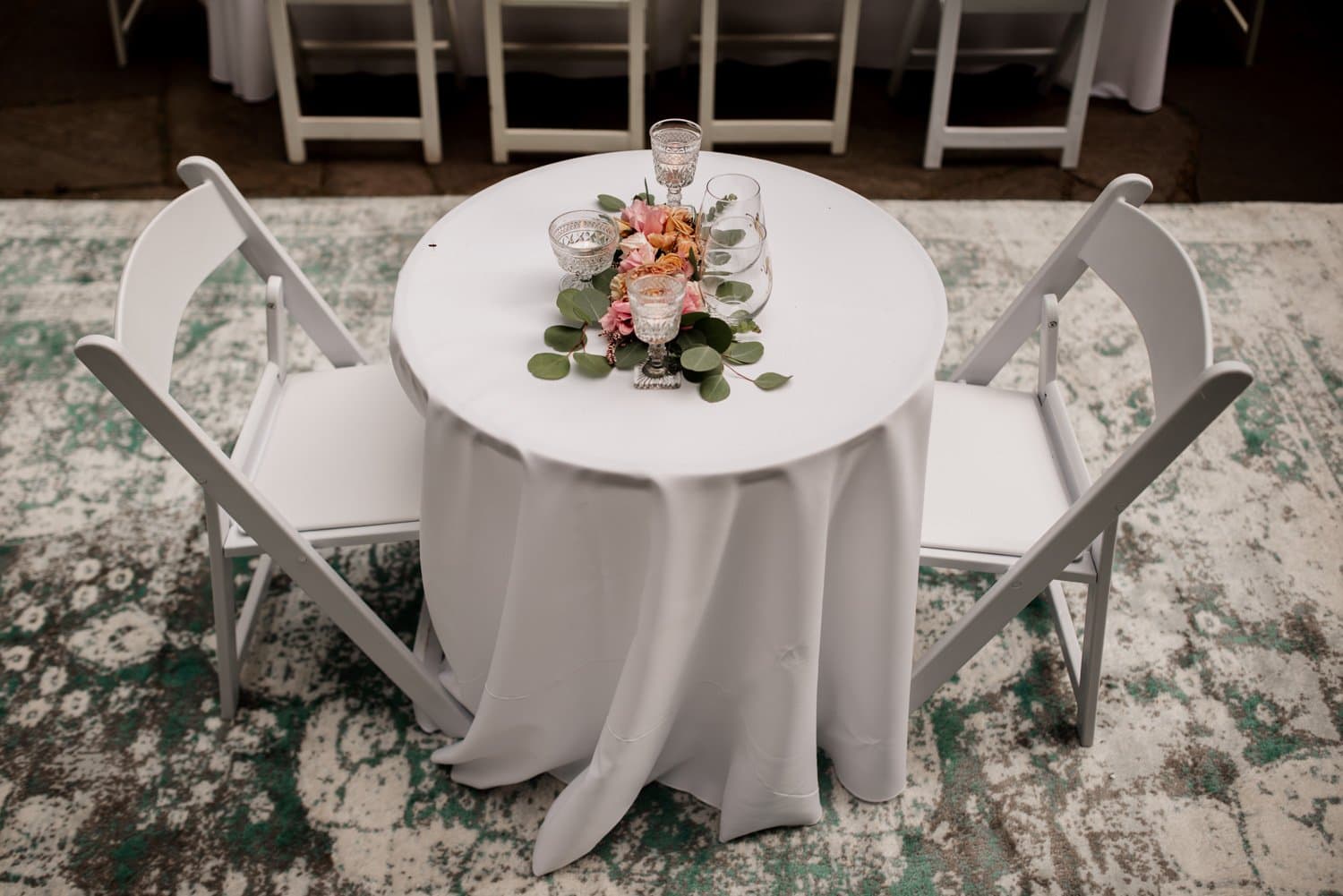 A round table with a white cloth and two white chairs, featuring a floral centerpiece and glasses, set on a patterned rug at Leach Botanical Garden.