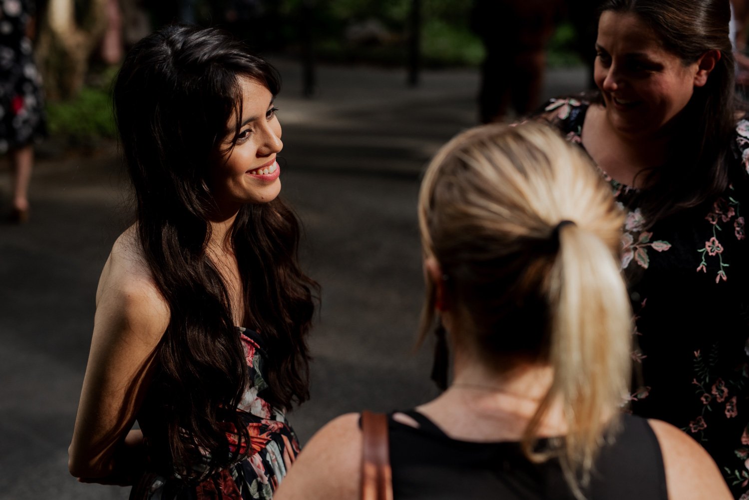 Three women sit together outdoors at Leach Botanical Garden, smiling and chatting in the sunlight. One woman has long dark hair, while the other two have lighter hair. They are enjoying a joyful moment surrounded by greenery.