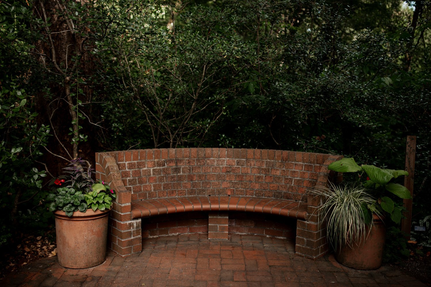 A curved brick bench is surrounded by potted plants and lush greenery in a garden setting.