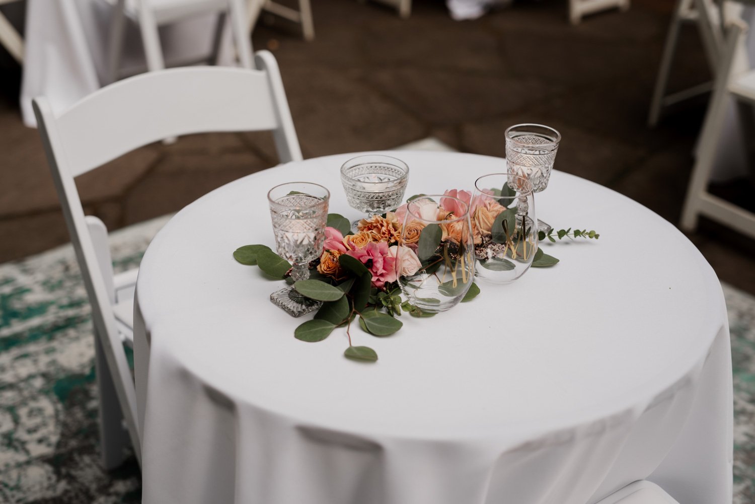 In Leach Botanical Garden, a white table with a floral centerpiece and crystal glassware is surrounded by white chairs, set up for a wedding.