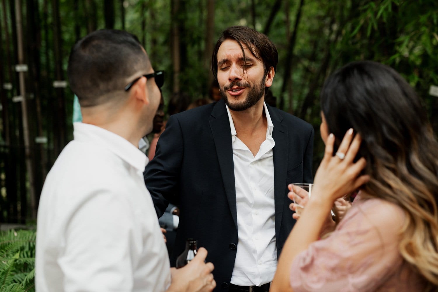 Three people stand amidst the lush greenery of Leach Botanical Garden, chatting and laughing. One person raises a drink in celebration during a garden wedding under the tree canopy.
