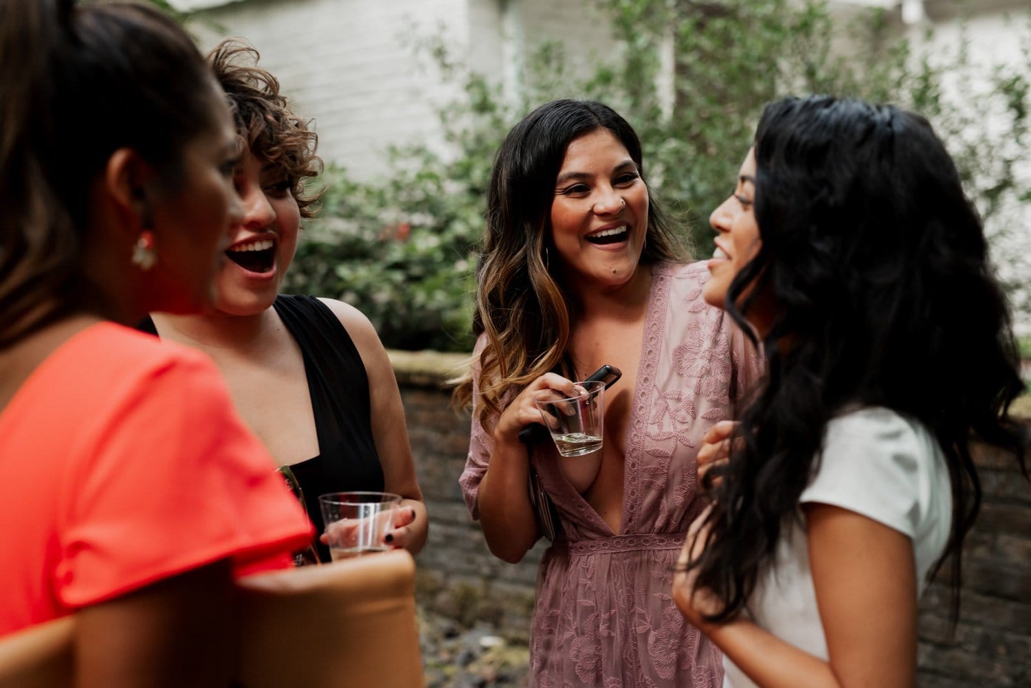 Four women laughing and chatting outdoors, holding drinks in a picturesque garden setting with vibrant flowers and greenery.