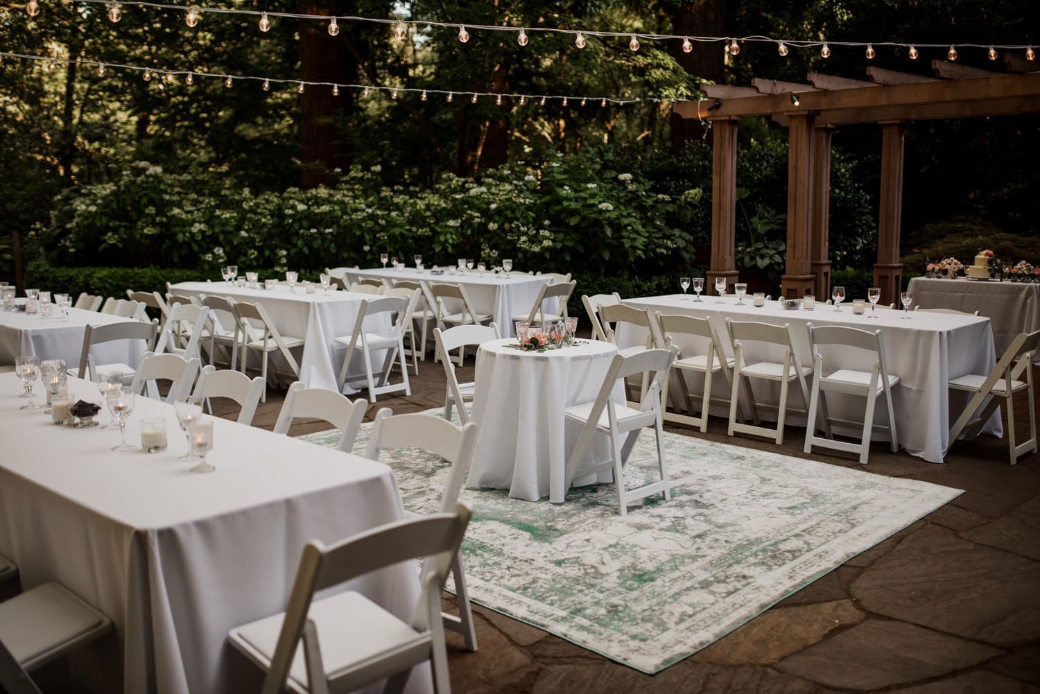 An outdoor wedding venue in Leach Botanical Garden with white tables and chairs, string lights hanging above, and a decorative rug on stone flooring beneath a rustic wooden pergola.