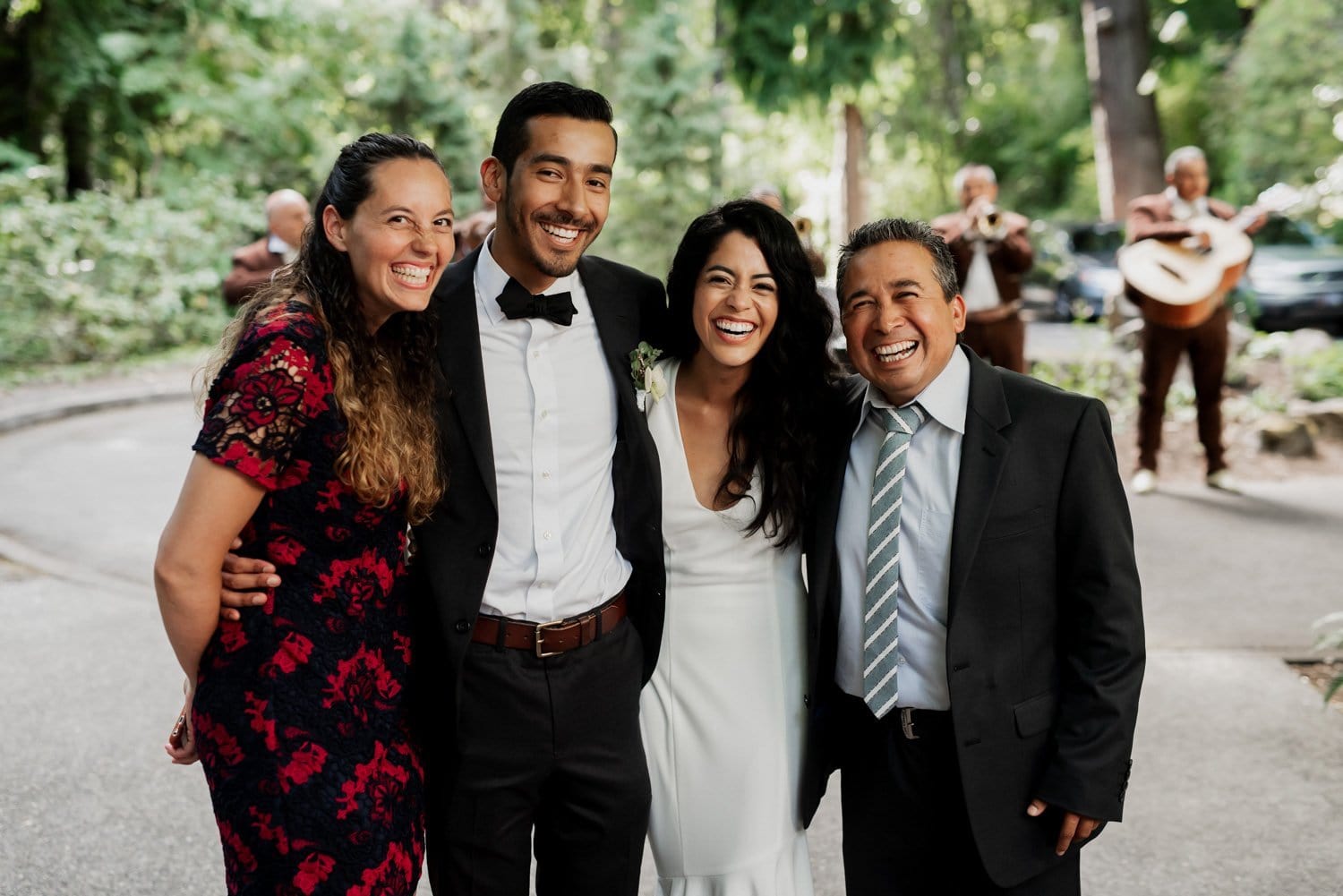 Four people in formal attire stand smiling outdoors with musicians playing in the background at a garden setting.
