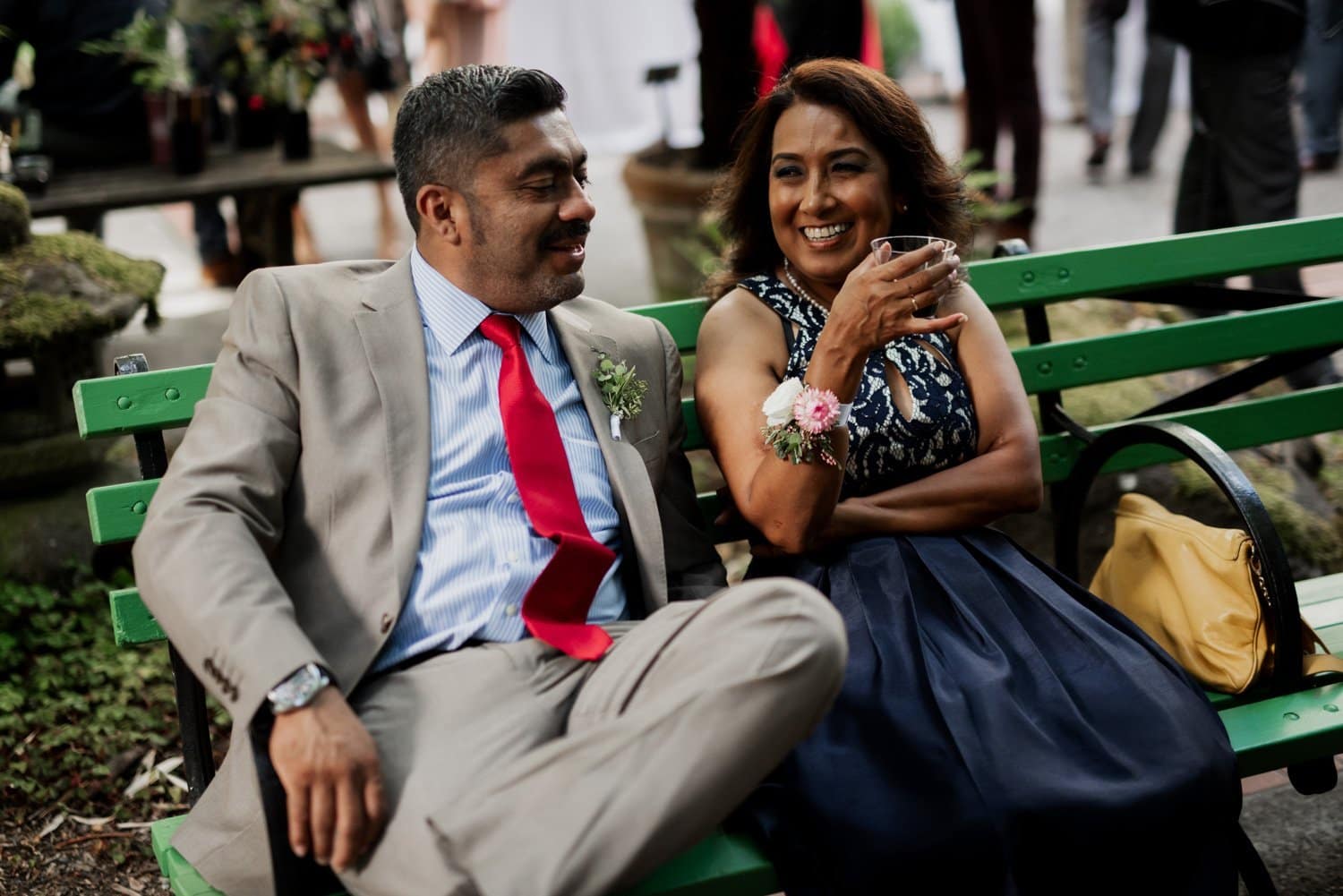 A man in a suit and a woman in a dress sit smiling on a green bench, with the vibrant setting of Leach Botanical Garden wedding events captured beautifully in the background.