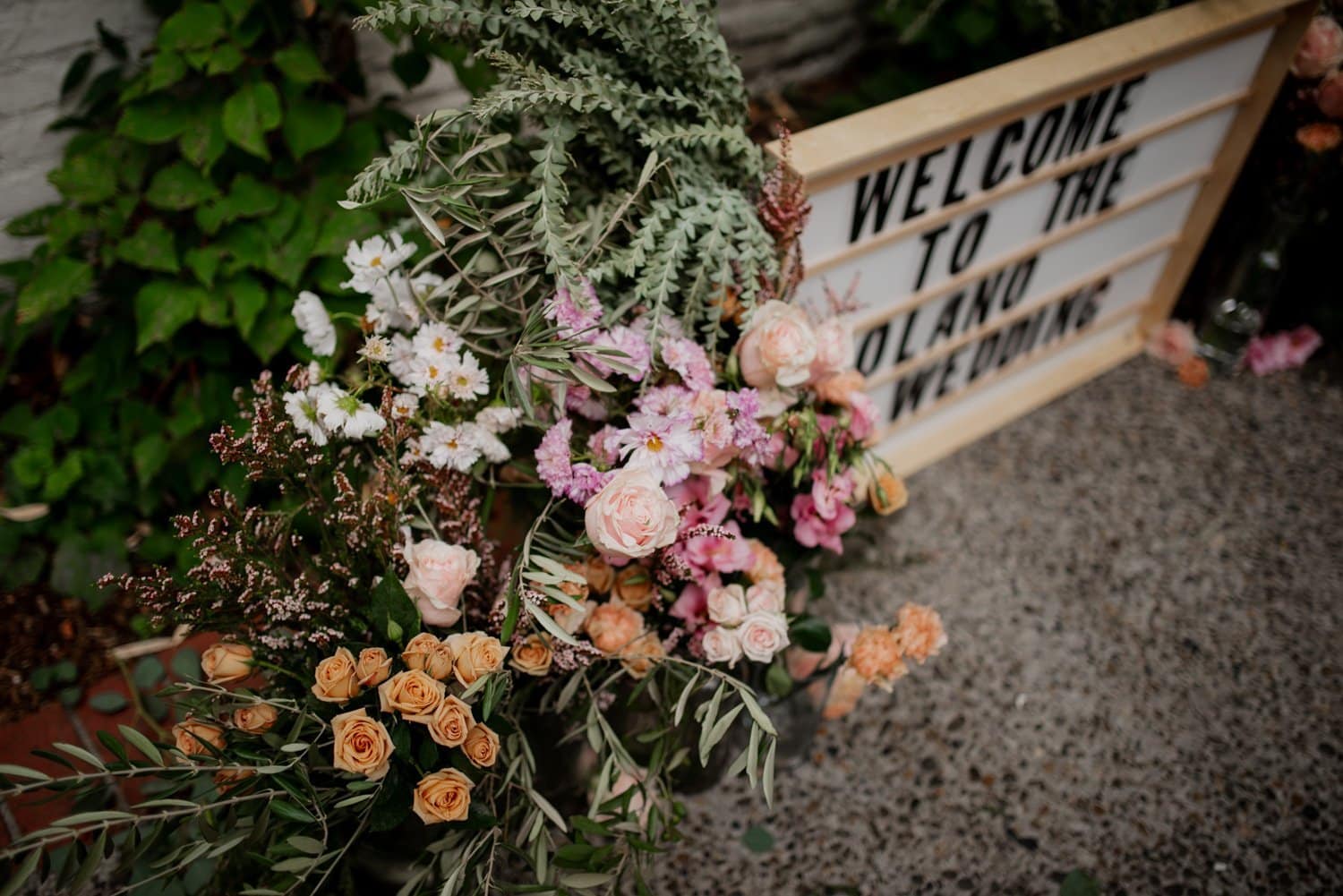 A floral arrangement of roses and greenery is displayed next to a sign with the words Welcome to the, suggesting a wedding at Leach Botanical Garden.