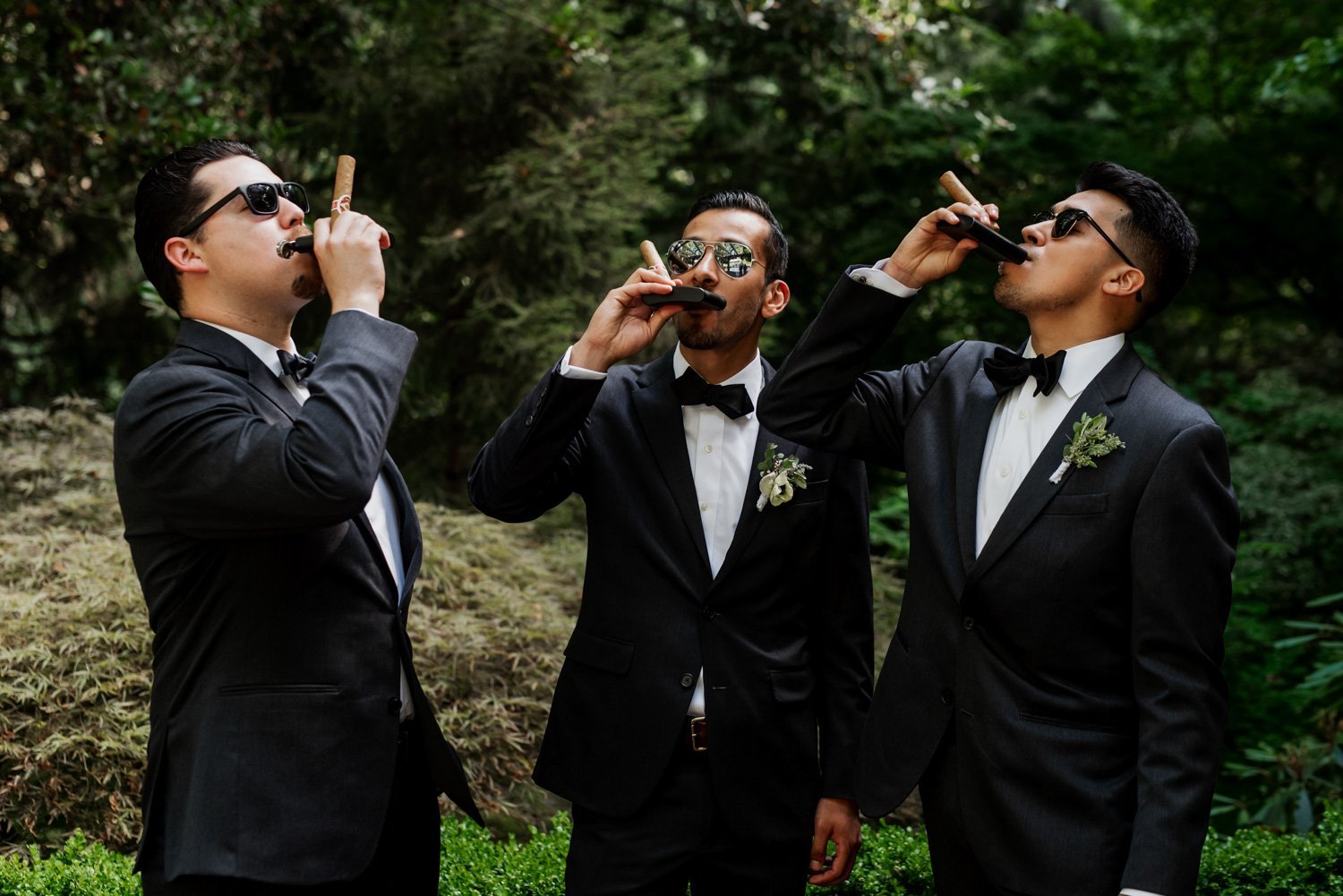 Three men in suits and sunglasses enjoy cigars surrounded by lush greenery at Leach Botanical Garden.