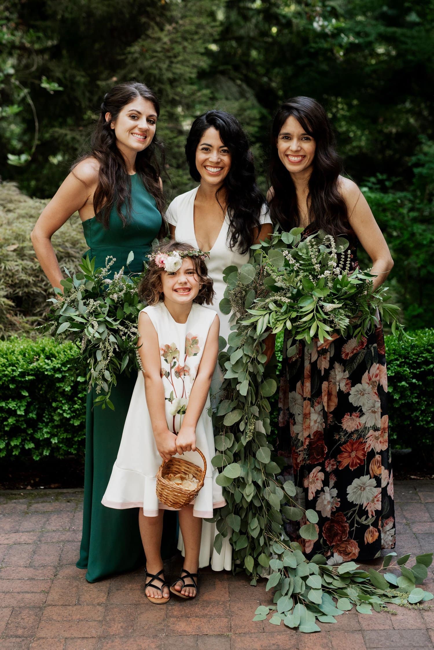 Four women stand together outdoors, surrounded by greenery. One woman holds a flower basket, and they are all smiling while wearing floral outfits at the Leach Botanical Garden wedding.