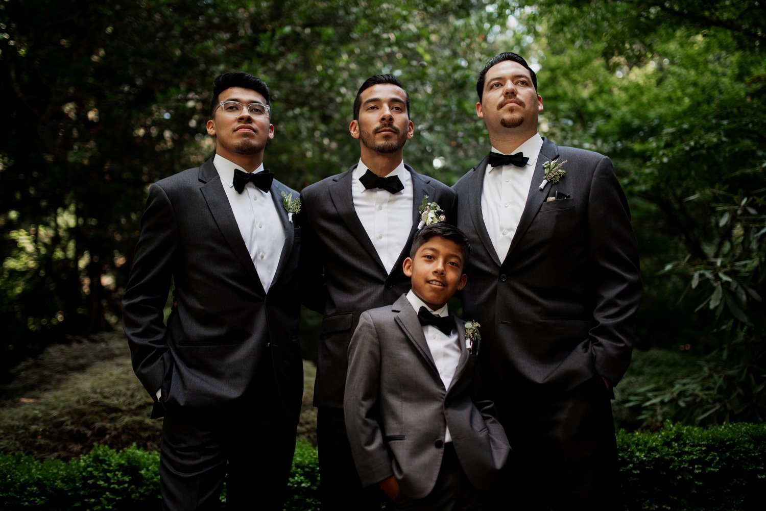 Four people in tuxedos stand together in a garden setting, with abundant green foliage visible in the background at Leach Botanical Garden.