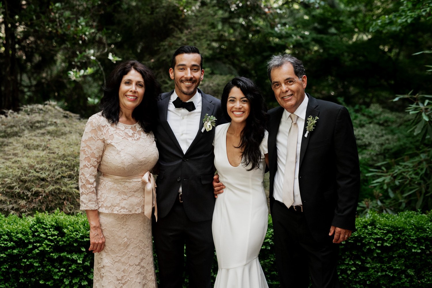 A bride and groom stand with two older adults, all smiling amidst the lush greenery and vibrant flowers at Leach Botanical Garden. The group is dressed elegantly, celebrating a wedding occasion in an outdoor setting.