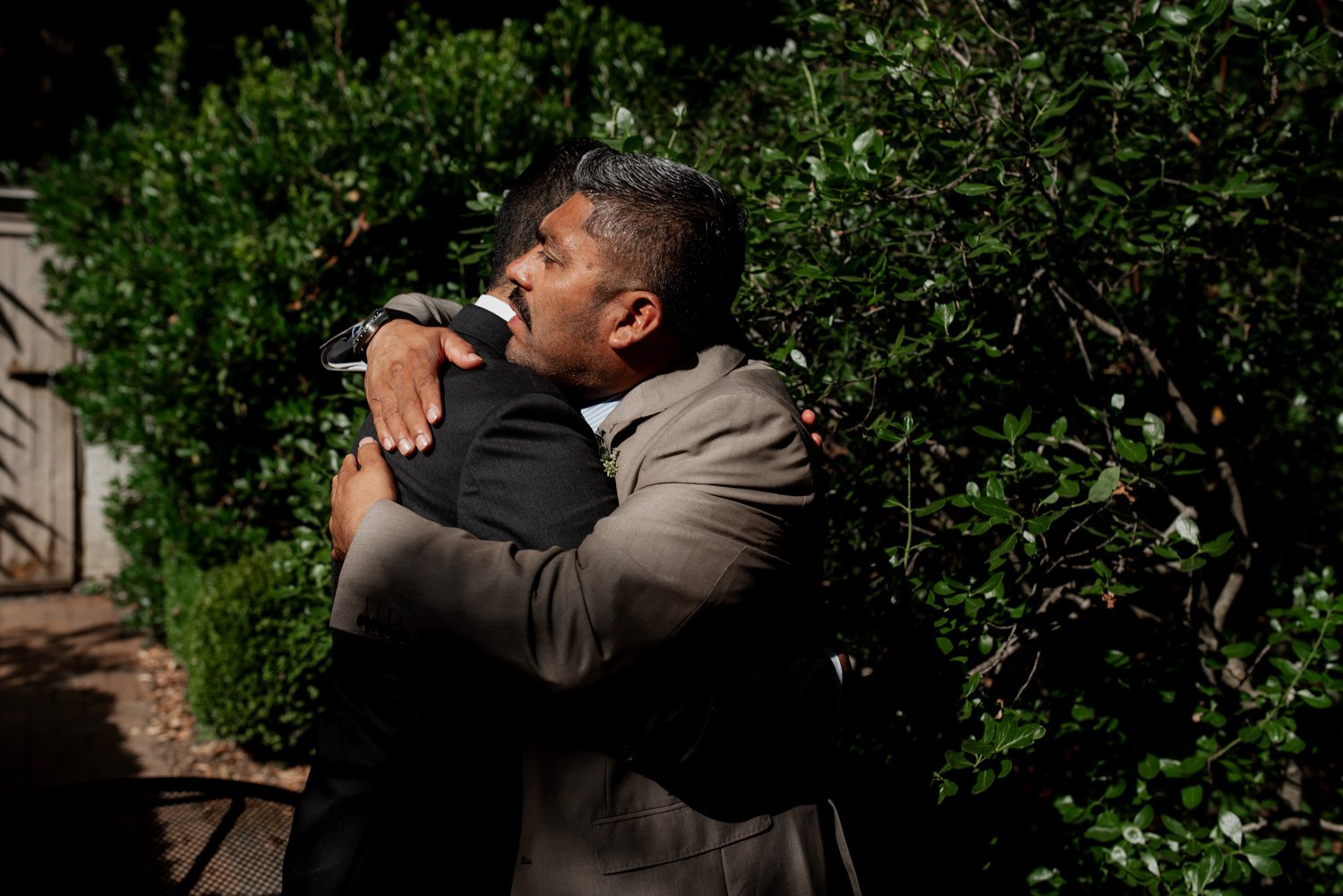 Two men in suits embrace warmly under the sun at Leach Botanical Garden, surrounded by lush green bushes.