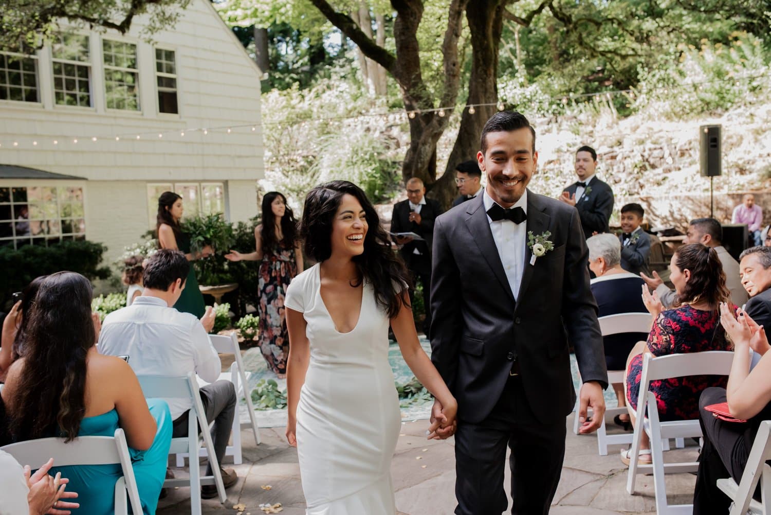 A couple in wedding attire holds hands and smiles while walking down the outdoor aisle at Leach Botanical Garden.