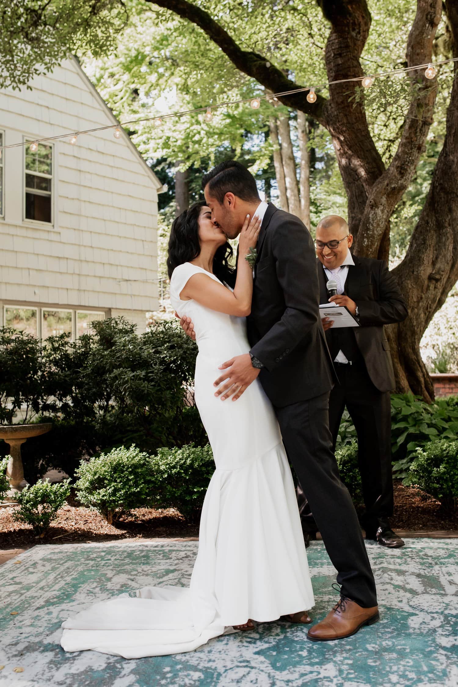 The bride and groom share a tender kiss beneath the lush canopy at their Leach Botanical Garden wedding, while the officiant smiles nearby under the trees.