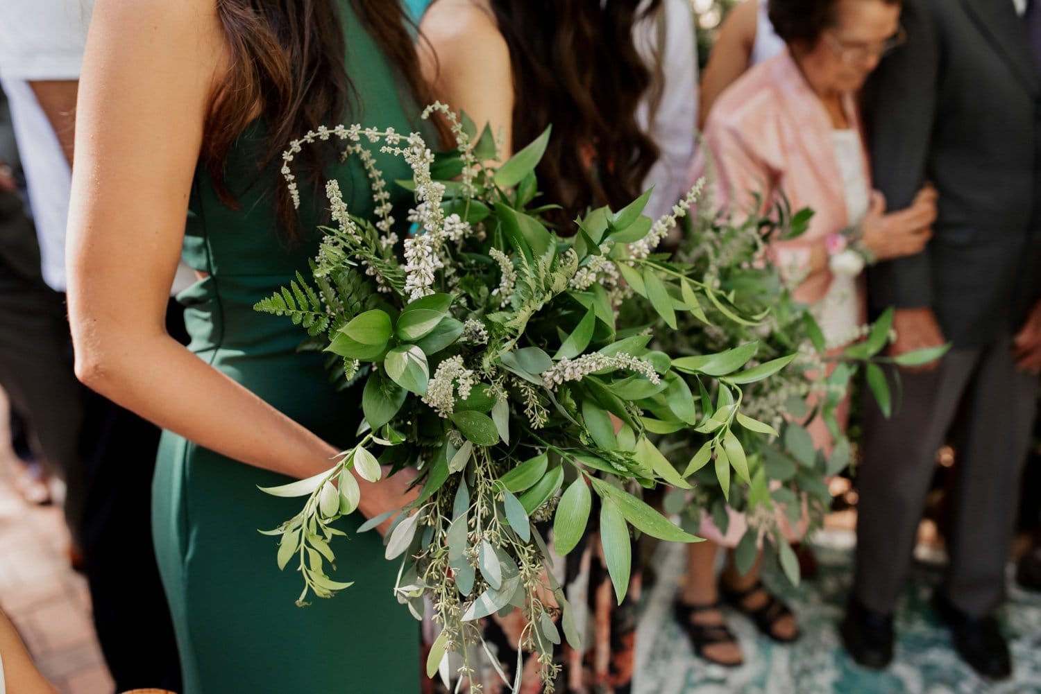 A person in a green dress holds a lush greenery bouquet at what seems to be a wedding at Leach Botanical Garden, with other attendees partially visible in the background.