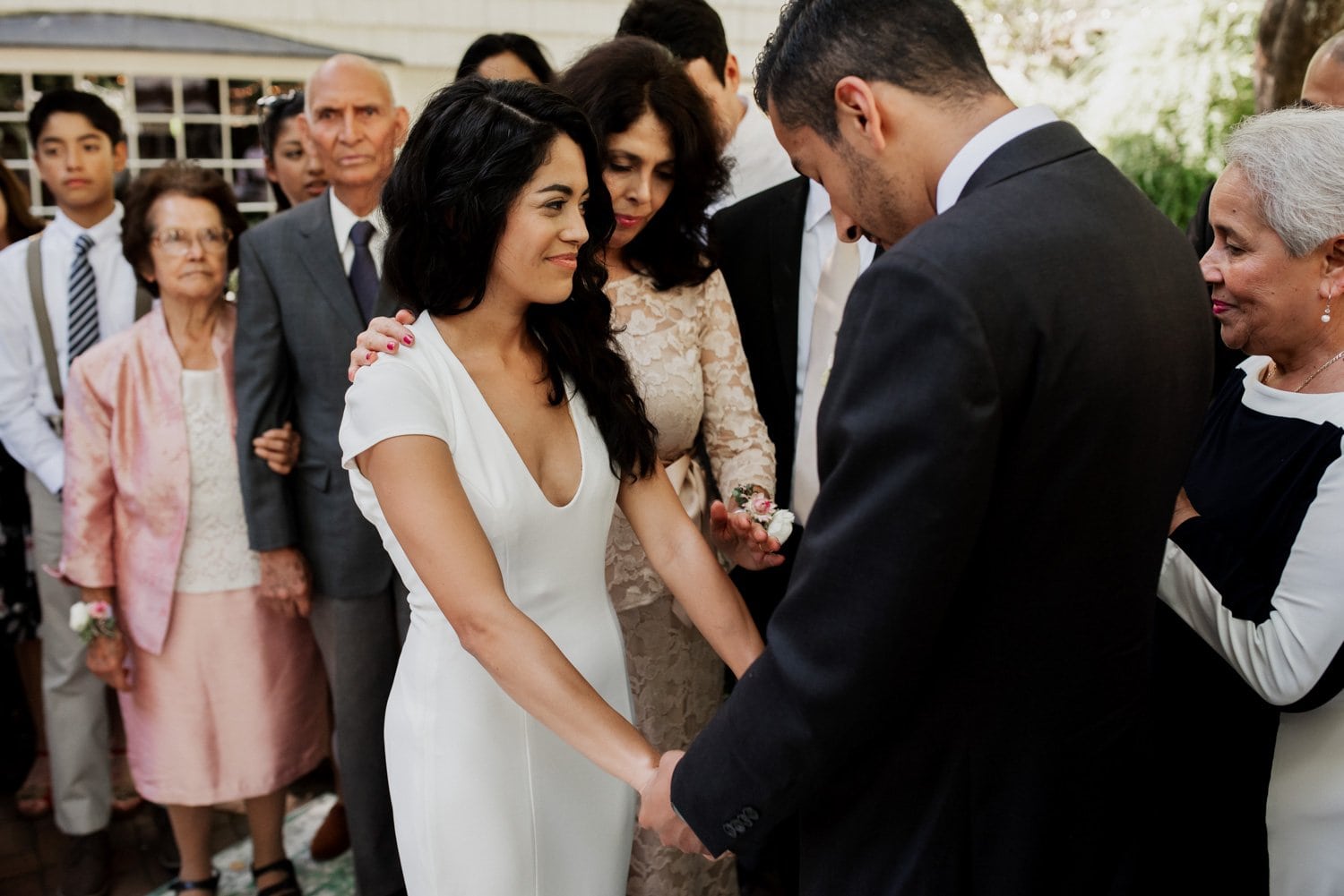 A bride and groom hold hands during their wedding ceremony at Leach Botanical Garden, with family members gathered around them.
