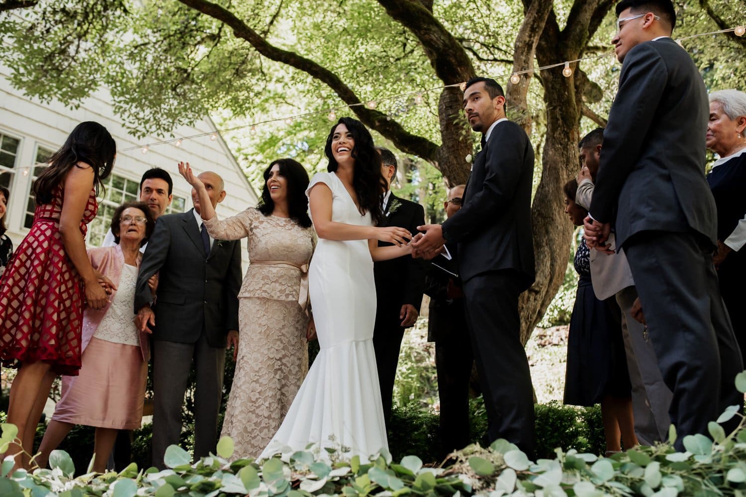 A couple holds hands during their outdoor ceremony at Leach Botanical Garden, surrounded by family and friends under a large tree.