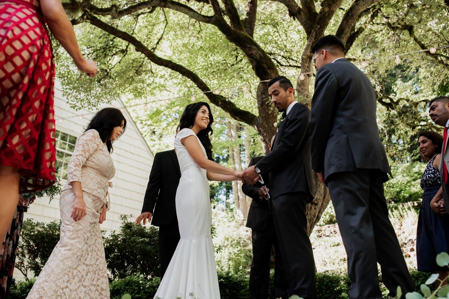 In the lush setting of Leach Botanical Garden, a couple stands together exchanging vows, surrounded by tall trees and their guests. A quaint house is visible in the background, enhancing the serene outdoor scene.
