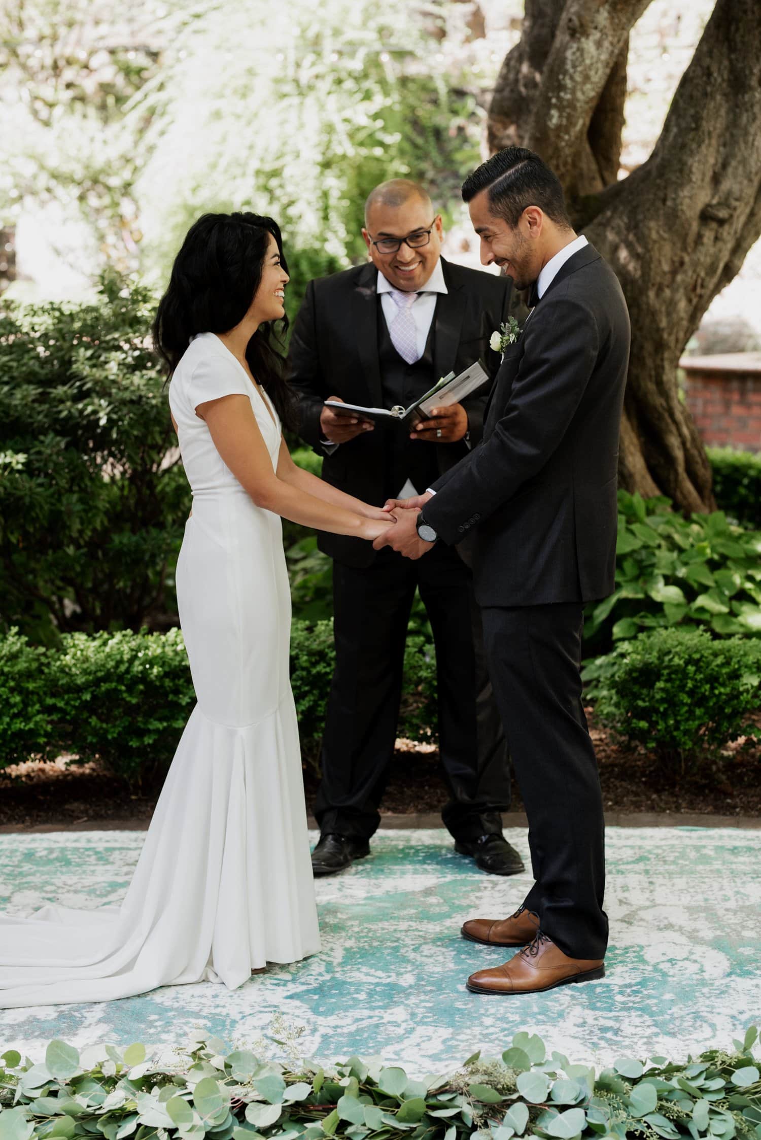 A couple holds hands during their outdoor wedding ceremony, standing under a large tree with an officiant at Leach Botanical Garden.