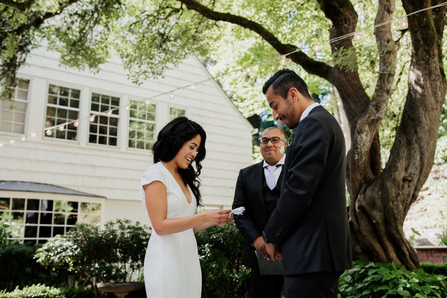 A bride reads her vows to the groom under a large tree at Leach Botanical Garden, with an officiant standing close by and a white building in the background.