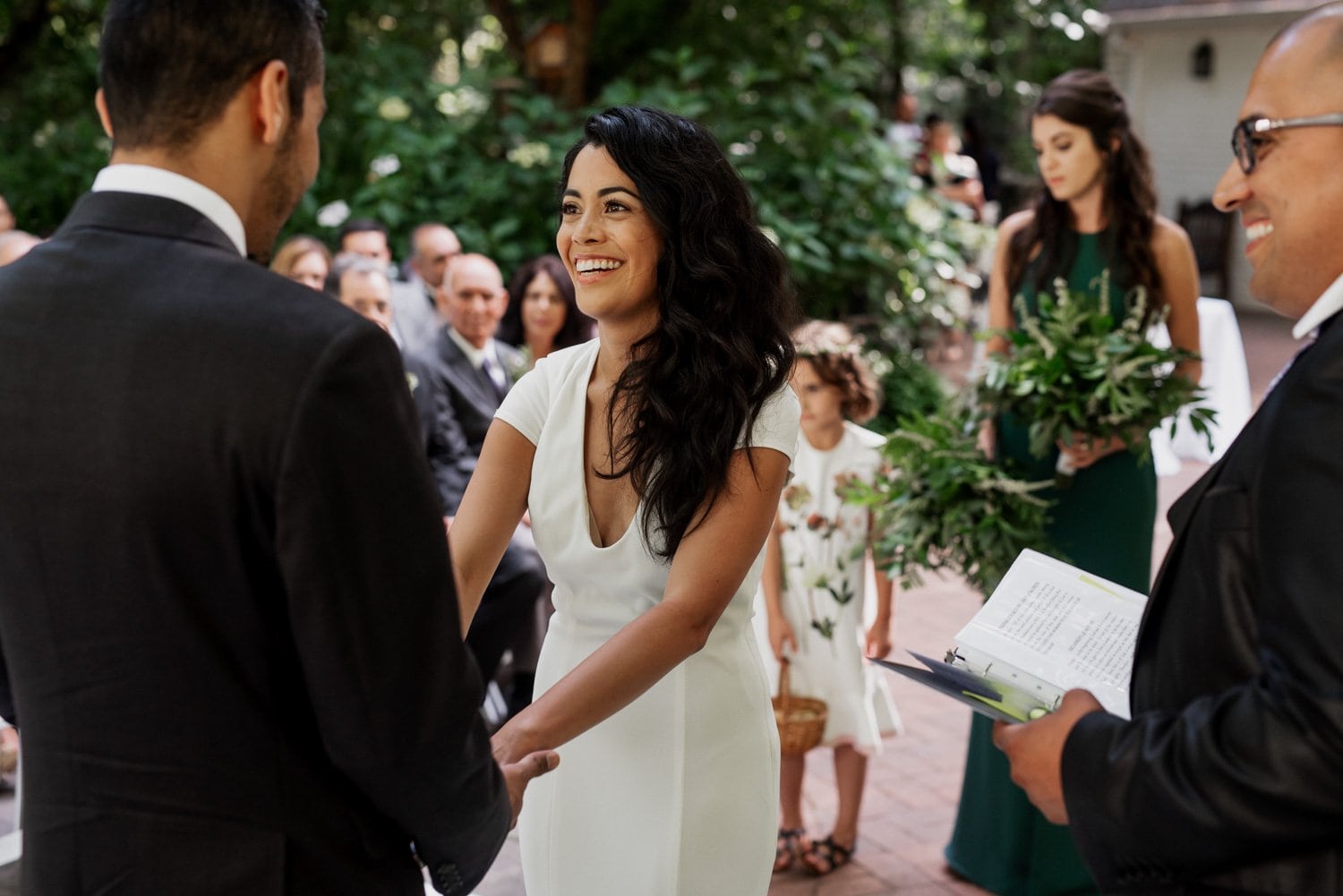 Bride smiling and holding hands with groom during an outdoor ceremony at Leach Botanical Garden, with guests seated behind.