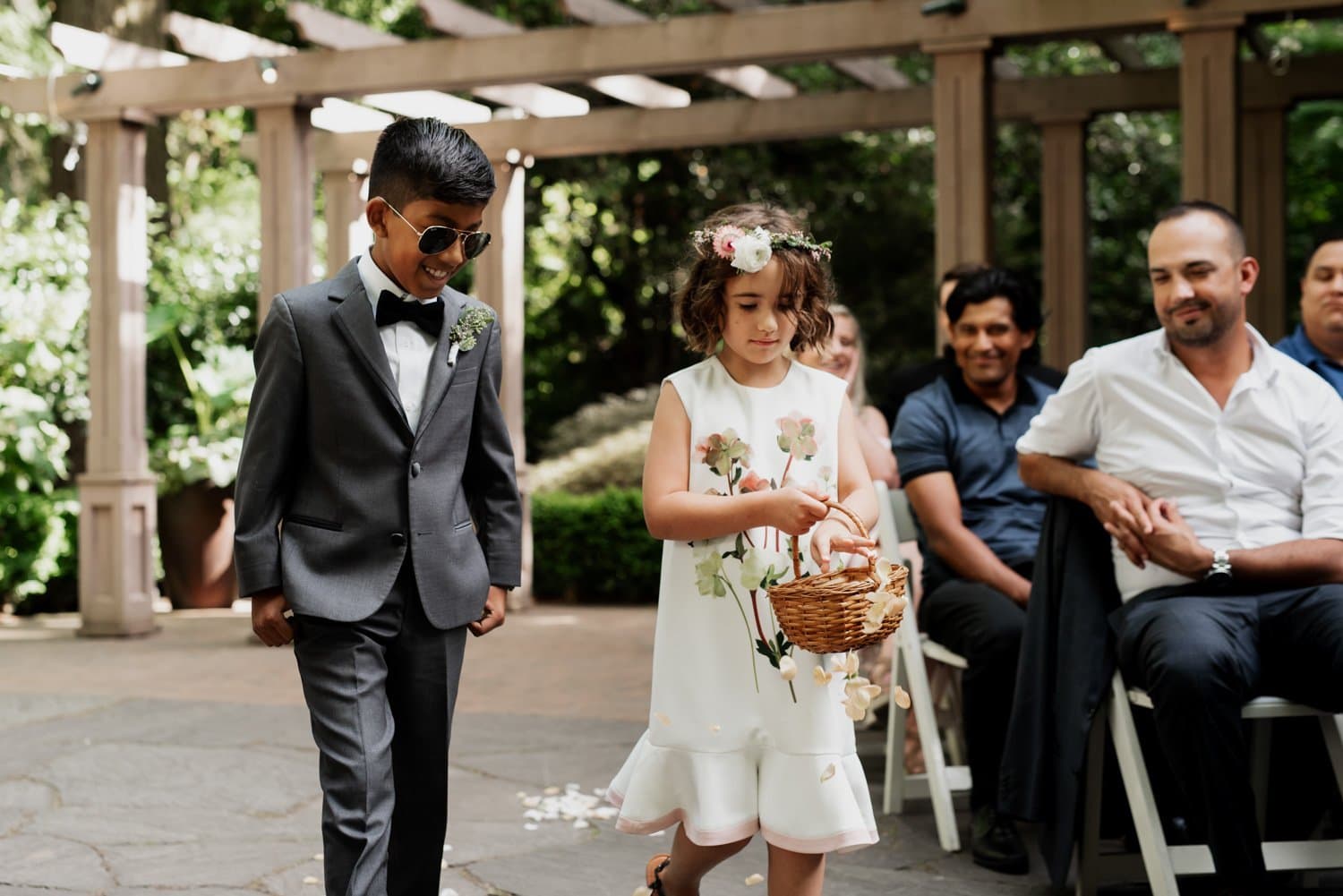 A boy in a suit and sunglasses walks next to a girl in a floral dress holding a basket at the Leach Botanical Garden wedding ceremony.