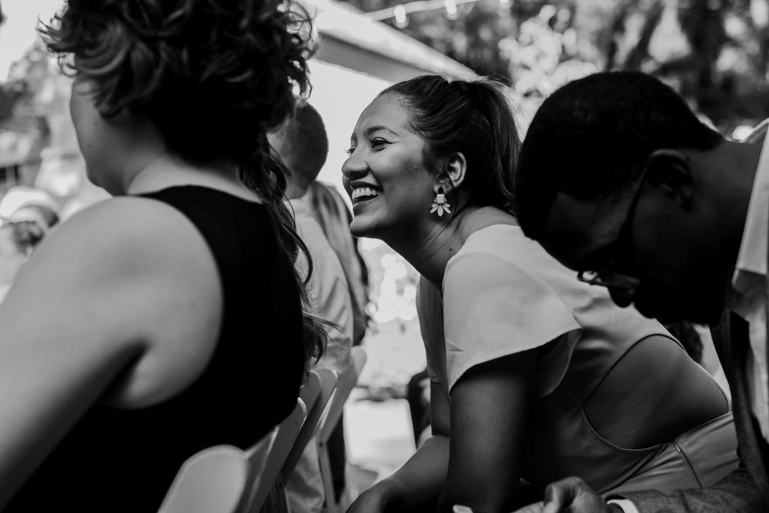 In black and white, a woman leans forward with a joyful smile among a lively seated group at an outdoor wedding at Leach Botanical Garden. The moment exudes timeless elegance and natural beauty.