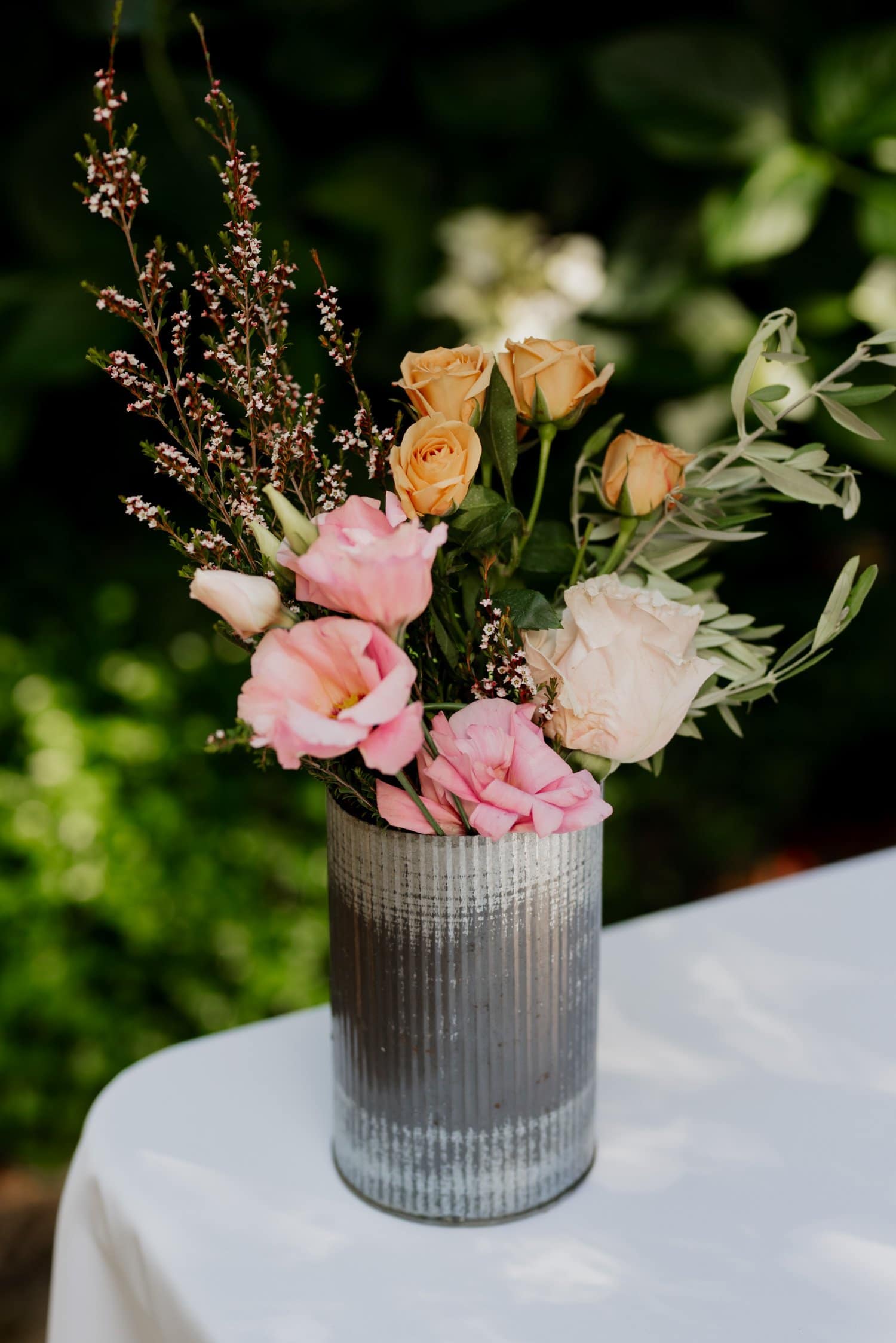 A silver vase filled with pink and peach roses surrounded by leafy greenery sits on a white table, with a blurred green background.