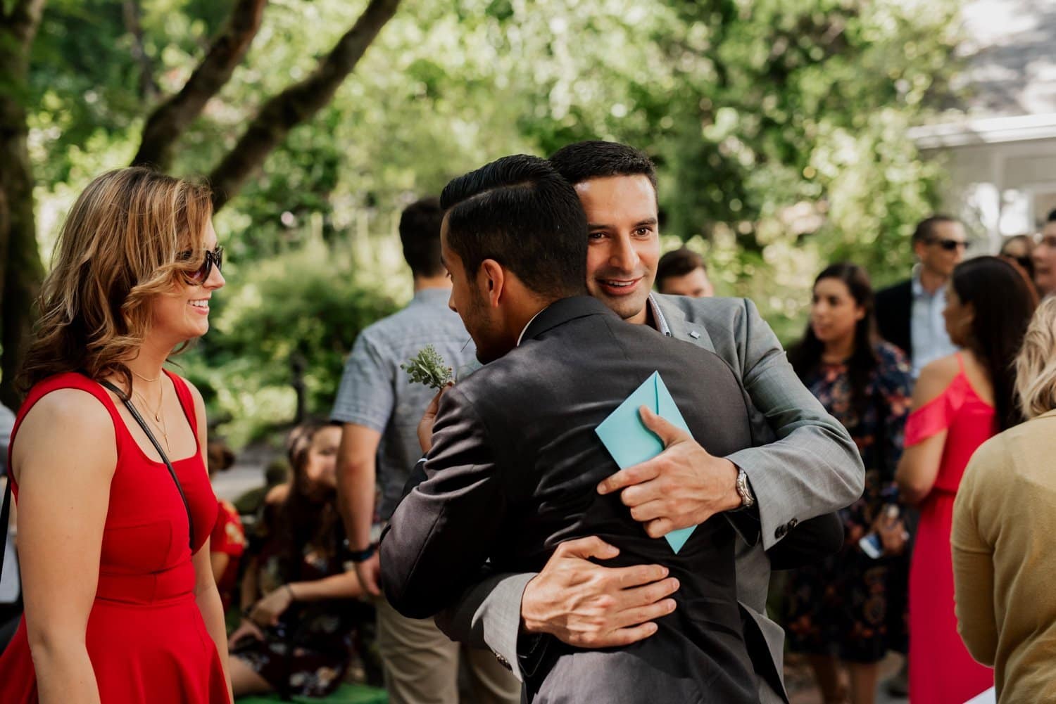 In the vibrant setting of Leach Botanical Garden, a group of formally dressed individuals is seen smiling and hugging, creating a joyful atmosphere during an outdoor event.