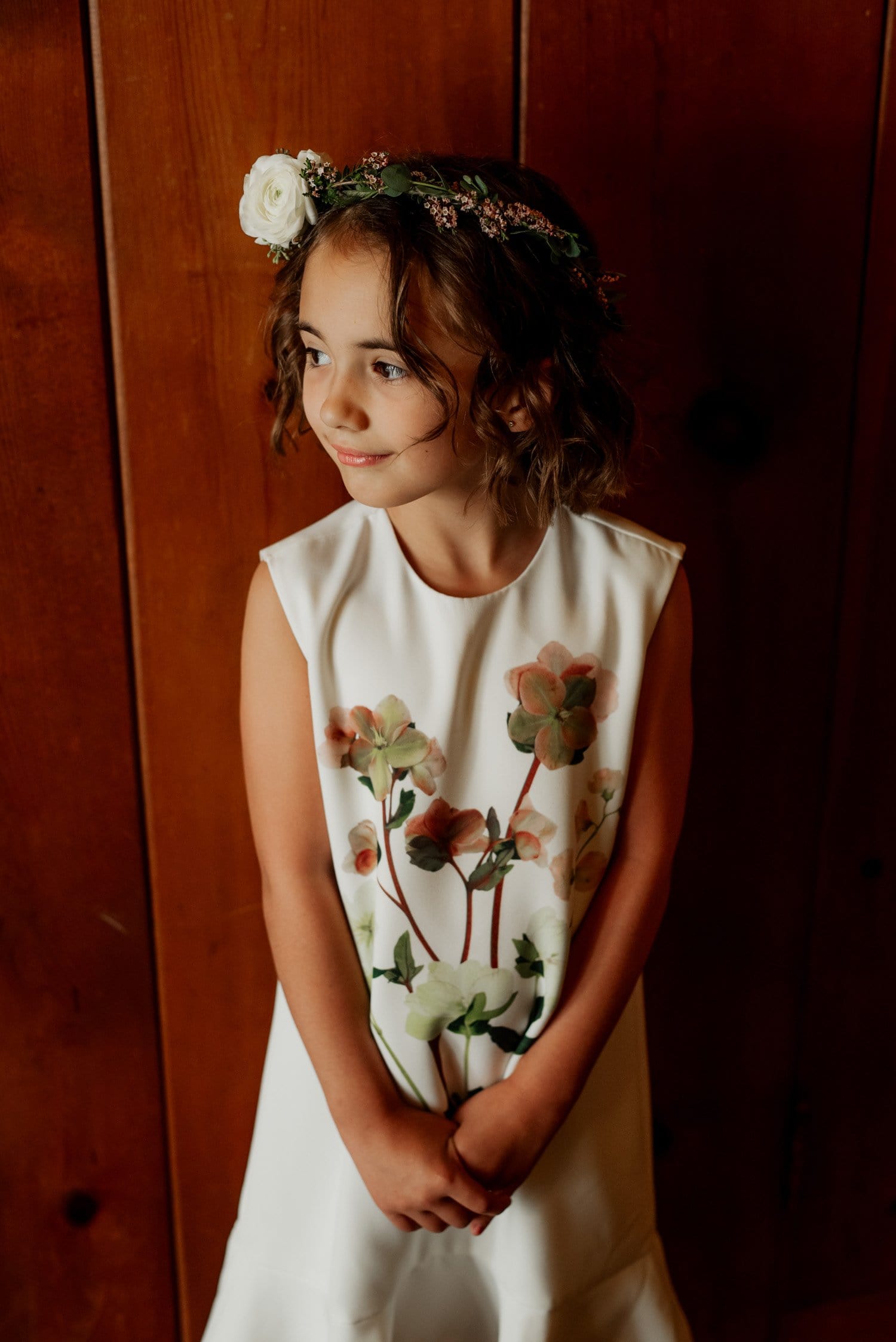 A young girl in a white floral dress and flower crown stands against a wooden wall, looking to the side at a wedding in Leach Botanical Garden.