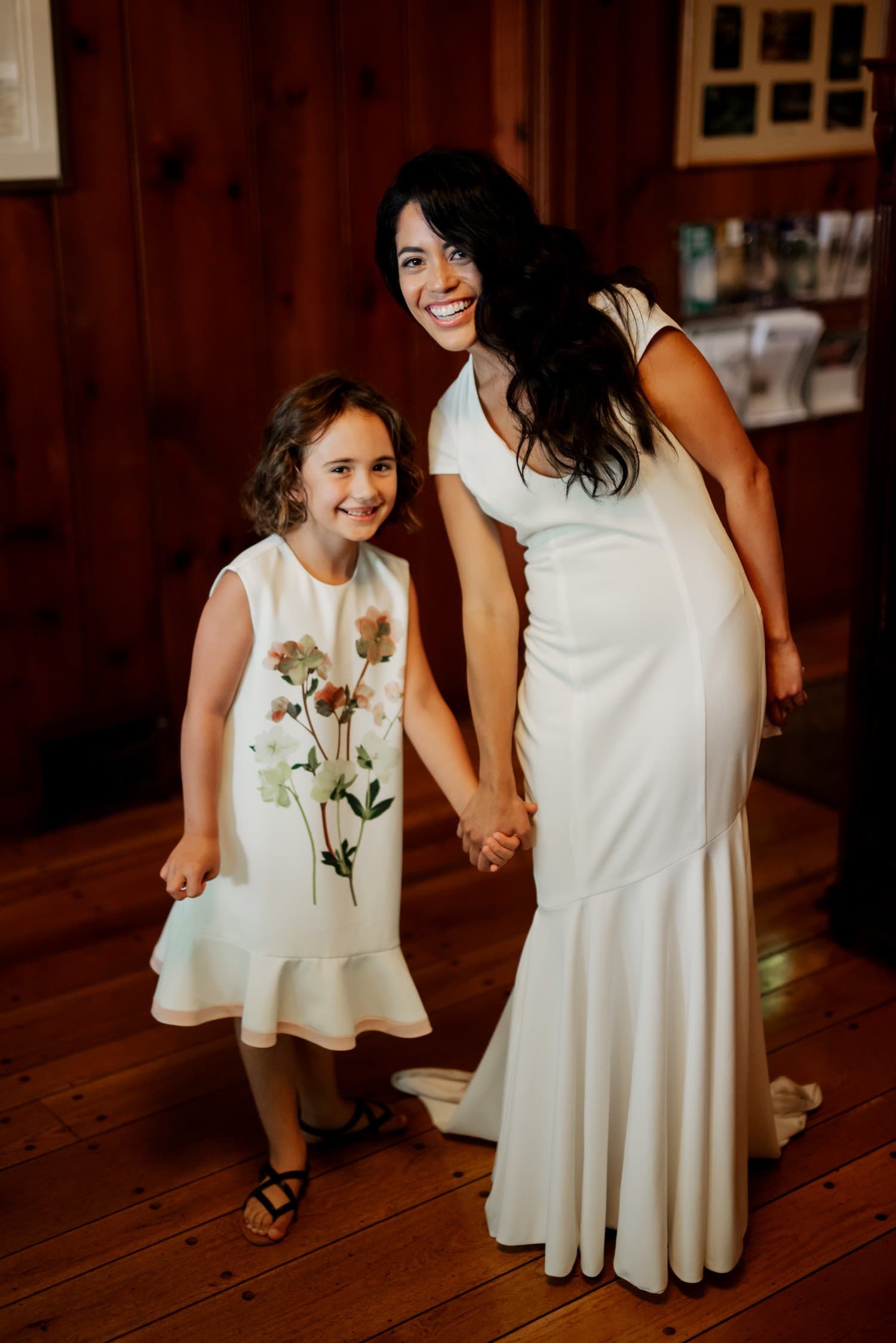 A woman in a white dress and a smiling girl in a floral dress hold hands on an elegant wooden floor indoors at Leach Botanical Garden.