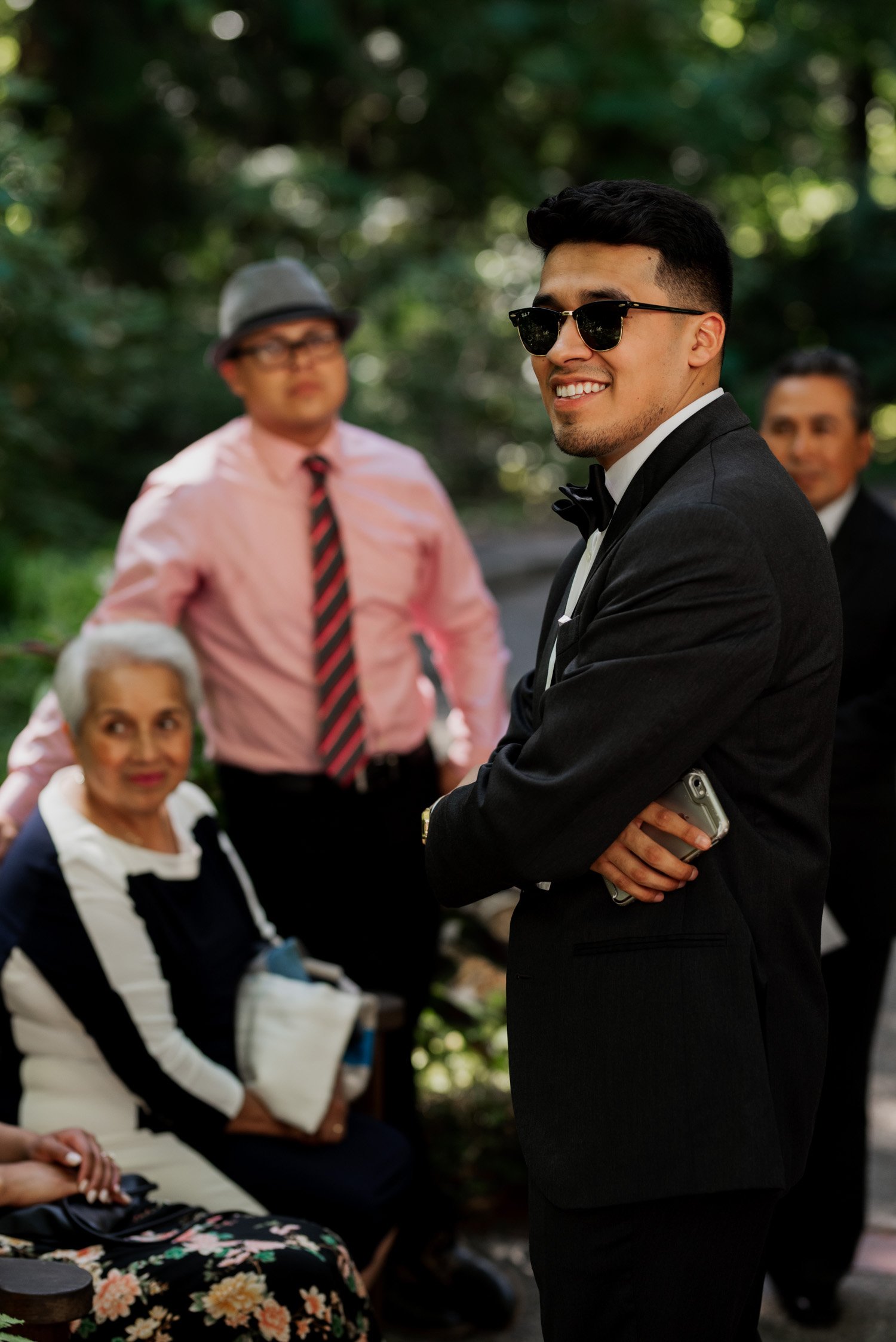 A man in a suit and sunglasses smiles while holding a phone at Leach Botanical Garden during a wedding. Three others are visible in the background amidst the lush outdoor scenery.