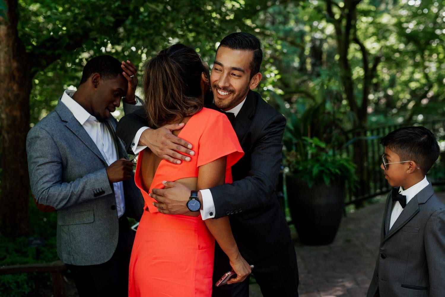 A man in a suit embraces a woman in an orange dress outdoors at Leach Botanical Garden, while two others, including a young boy in a tuxedo, observe the moment.