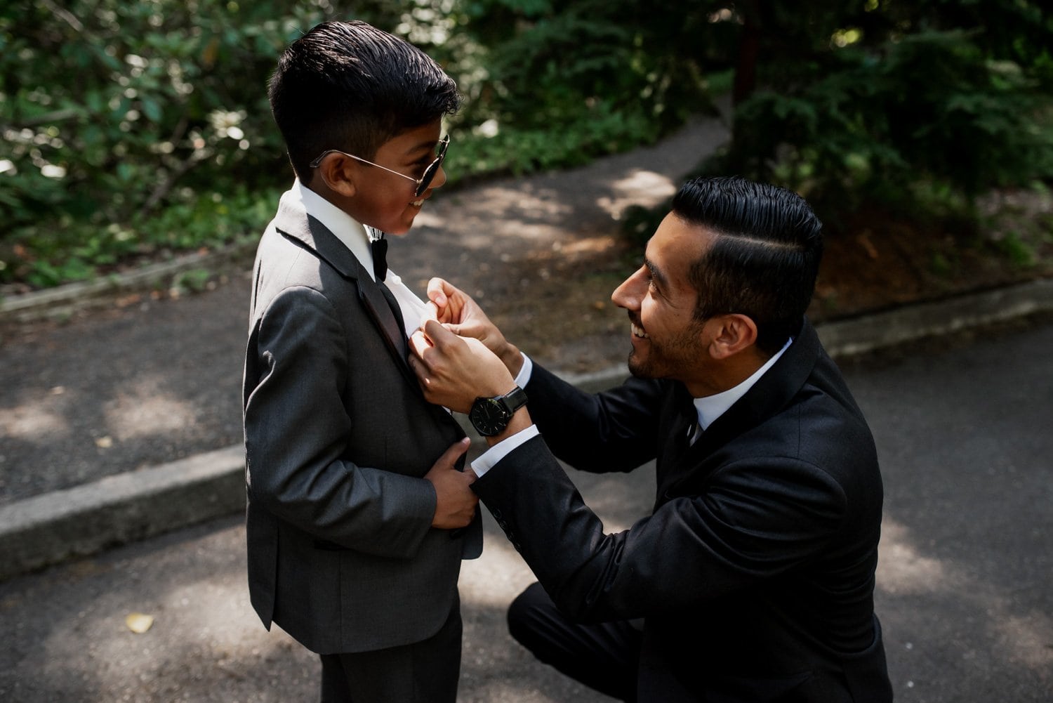 In the scenic Leach Botanical Garden, a man in an elegant suit adjusts a young boys tie outdoors, both smiling warmly. The setting and their attire suggest they are part of a wedding celebration.