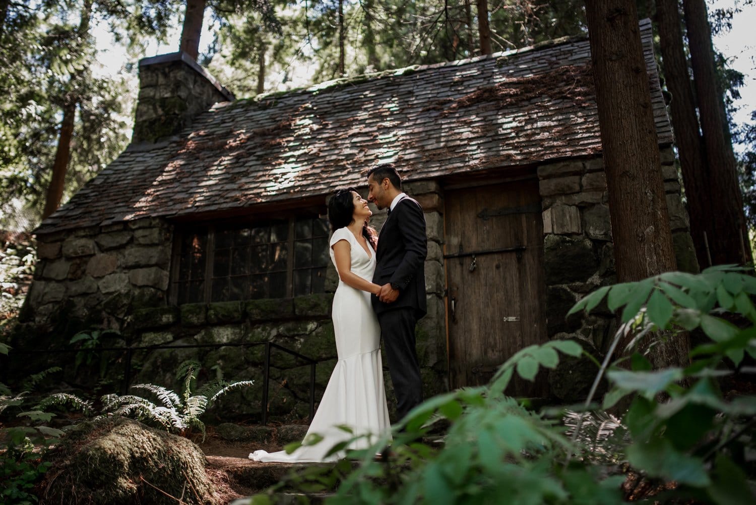 Bride and groom hold hands, gazing at each other in front of a rustic stone cabin surrounded by the lush greenery and vibrant flowers of Leach Botanical Garden.