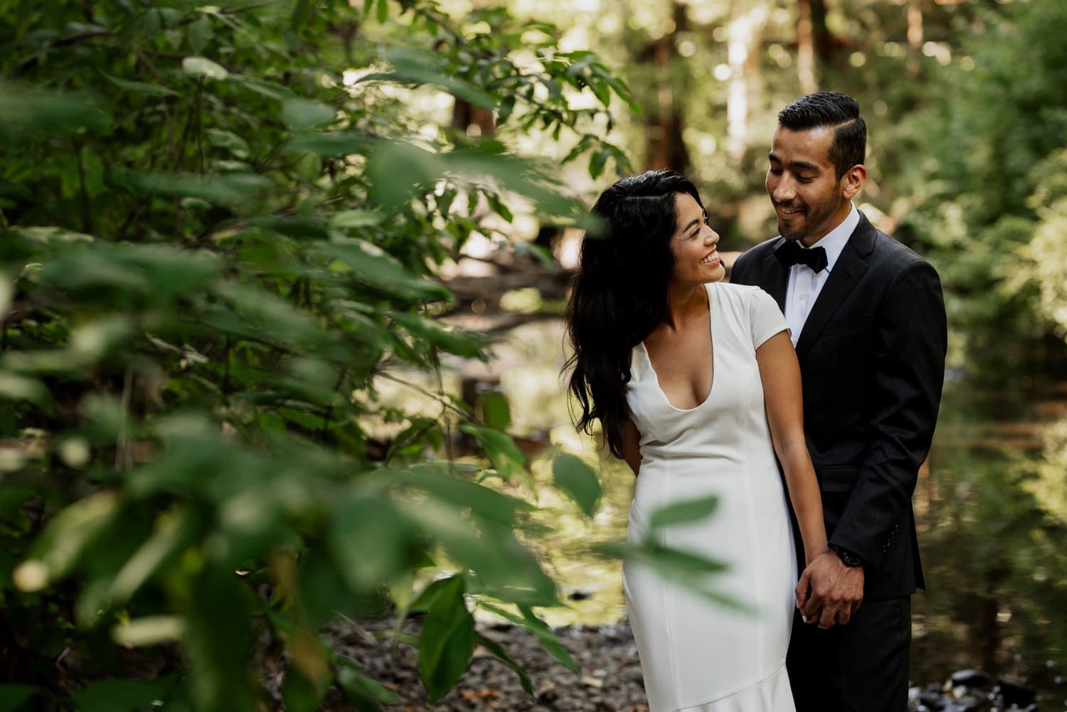 A couple smiling at each other by a forest stream, with the woman wearing a white dress and the man in a black suit, during their wedding at Leach Botanical Garden.