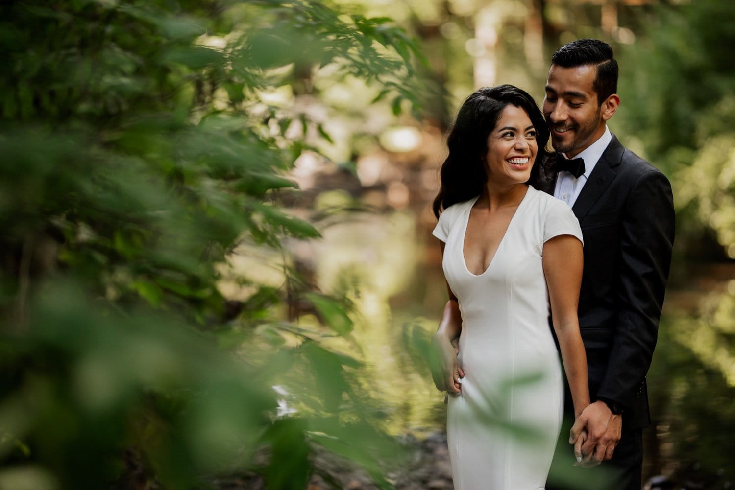 A couple in wedding attire smiles while standing together in the lush, green surroundings of Leach Botanical Garden.