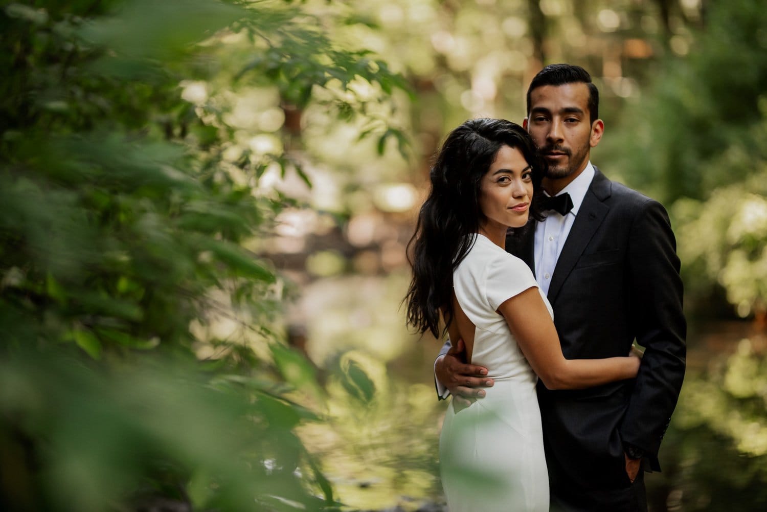 A couple embraces in a lush, green outdoor setting at Leach Botanical Garden. The man wears a suit, and the woman is in a white dress, highlighting their wedding day.