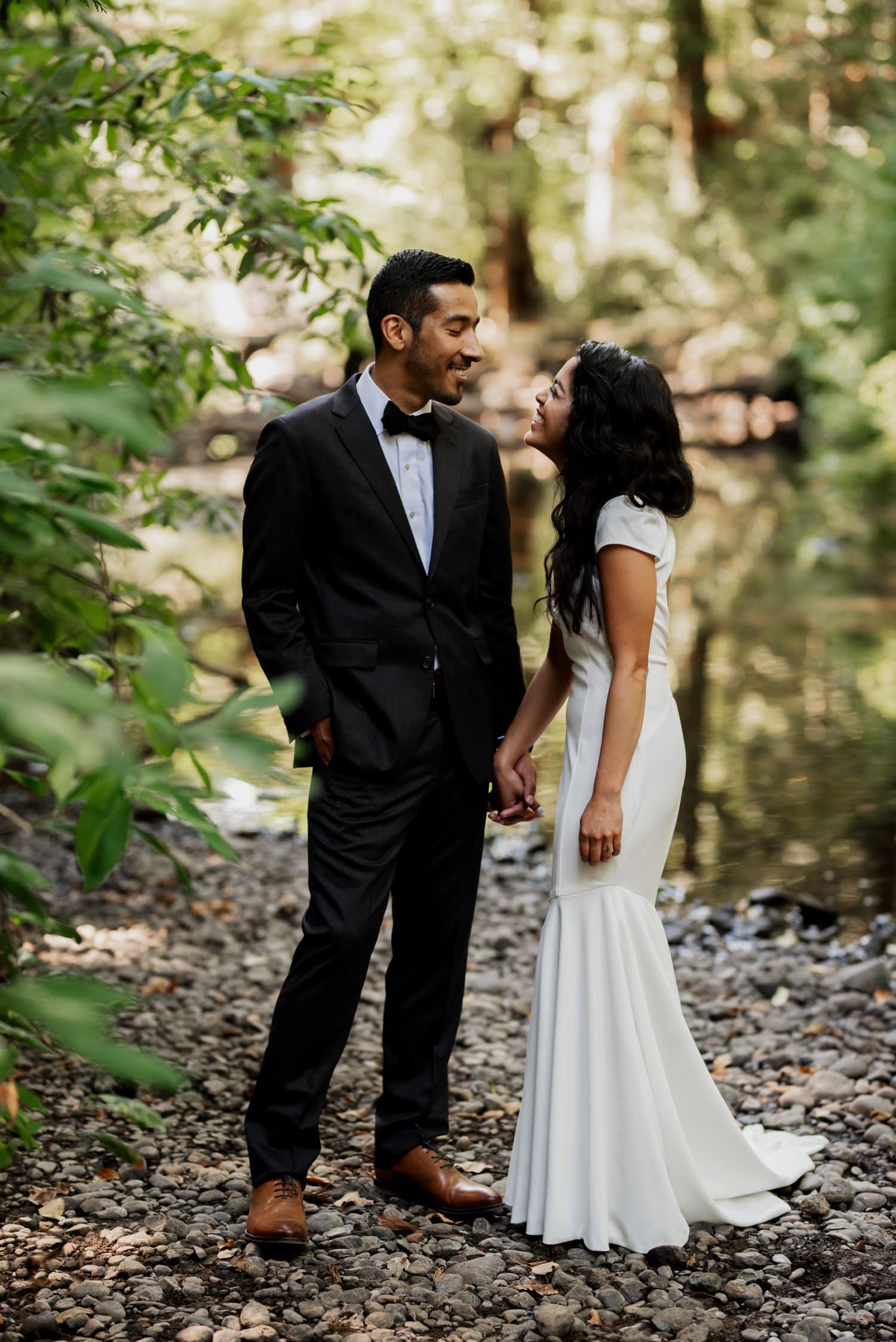 A couple stands on a rocky path by a river at Leach Botanical Garden, holding hands and smiling at each other, with lush greenery surrounding them.